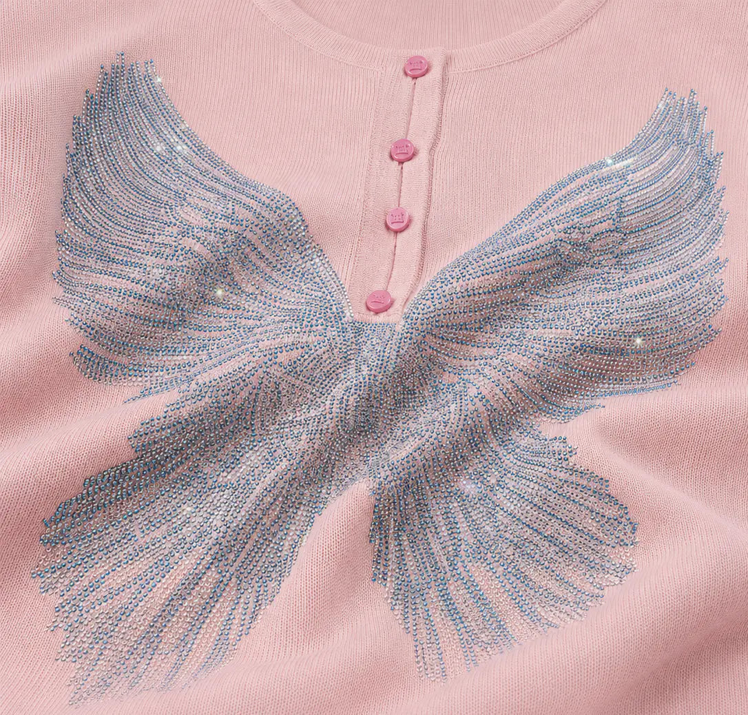 Close-up of a light pink knit top with a large sequined angel wing design across the chest and three pink buttons at the neckline. TCH Blue Rhinestone Angel Wings Cropped Hoodie