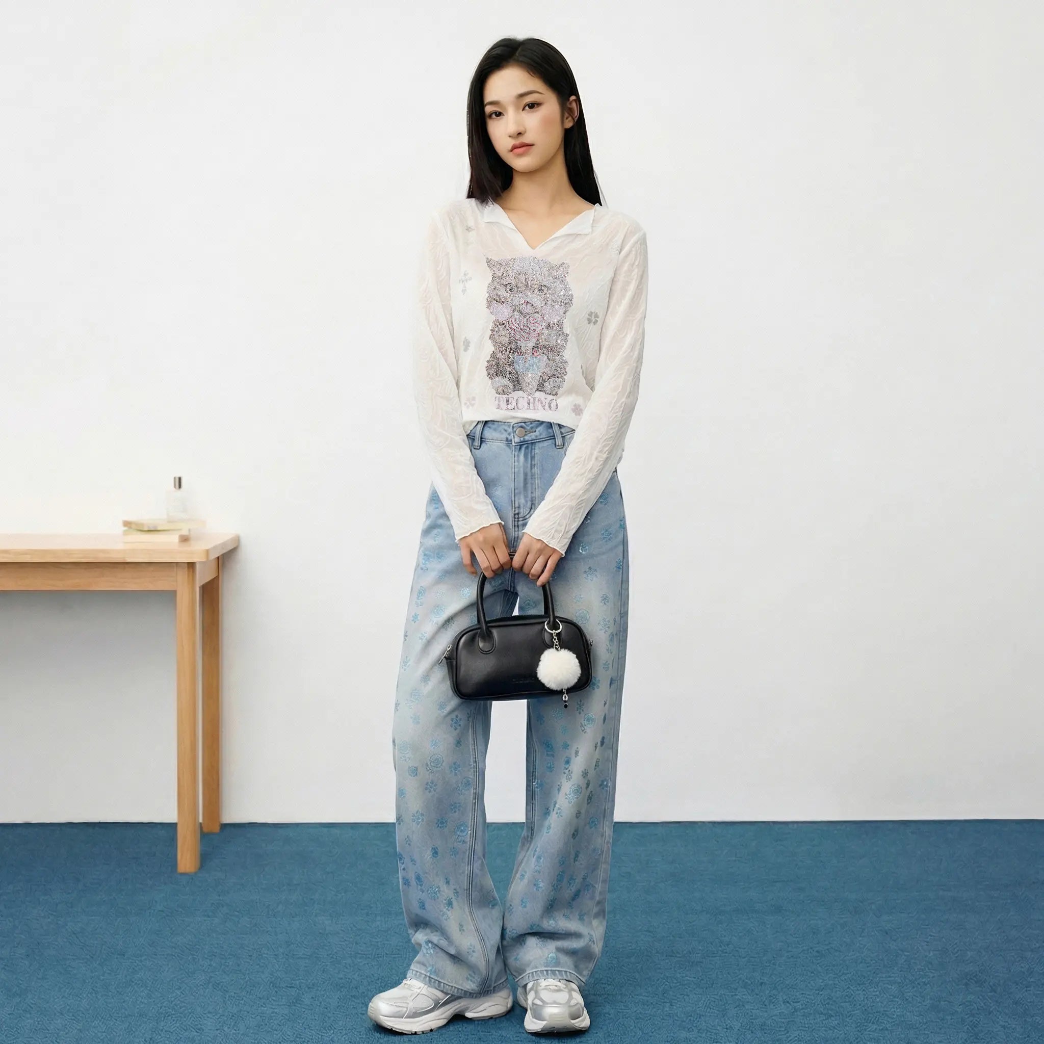 Woman wearing a white sweater with a graphic design and blue jeans, holding a black handbag with a pom-pom, against a plain background. TCH Blue Snowflake Rhinestone Denim Jeans