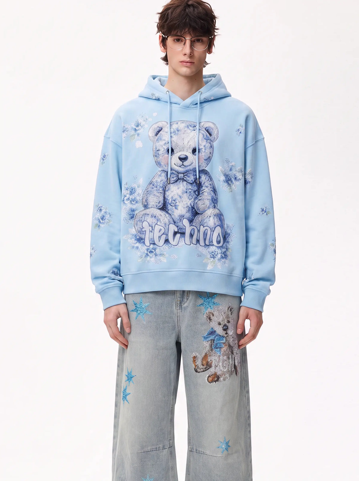 A model wearing a light blue hoodie with a large teddy bear graphic and the word 'felino' on the front, paired with light gray jeans featuring blue star prints and a teddy bear patch. TCH Blue Teddy Bear Floral Rhinestone Hoodie