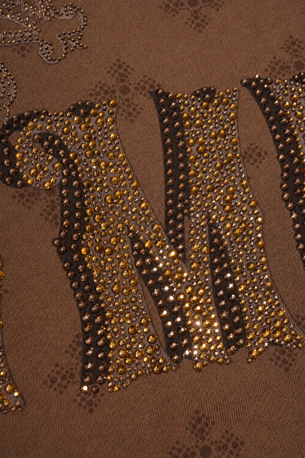 Close-up of a brown fabric surface with large decorative letters outlined in dark trim and densely covered with gold and brown rhinestones. TCH Brown Rhinestone Paisley Zip Up Hoodie