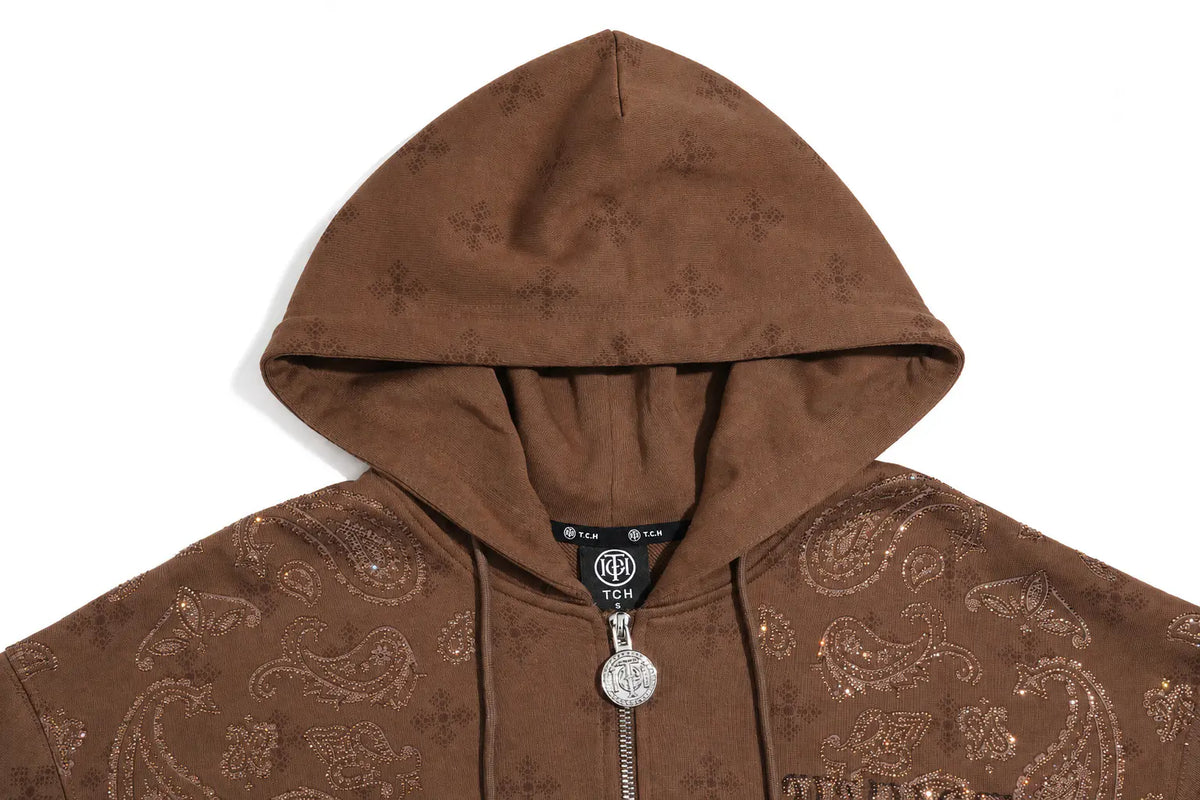 A close-up of a brown zip-up hoodie featuring an all-over paisley pattern, a silver-tone zipper with a round medallion pull, and a black neck label. TCH Brown Rhinestone Paisley Zip Up Hoodie
