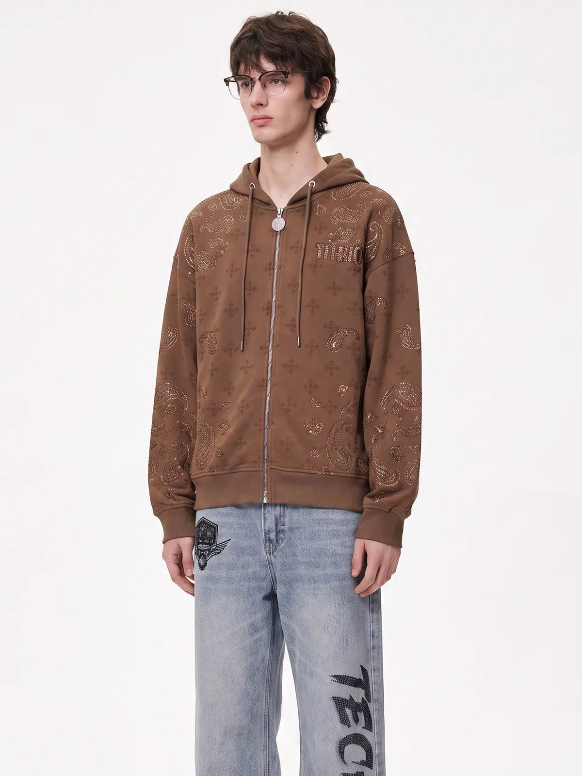A model wearing a brown zip-up hoodie with an all-over tonal abstract pattern and light-wash blue jeans with black graphic text, standing against a plain white studio background. TCH Brown Rhinestone Paisley Zip Up Hoodie