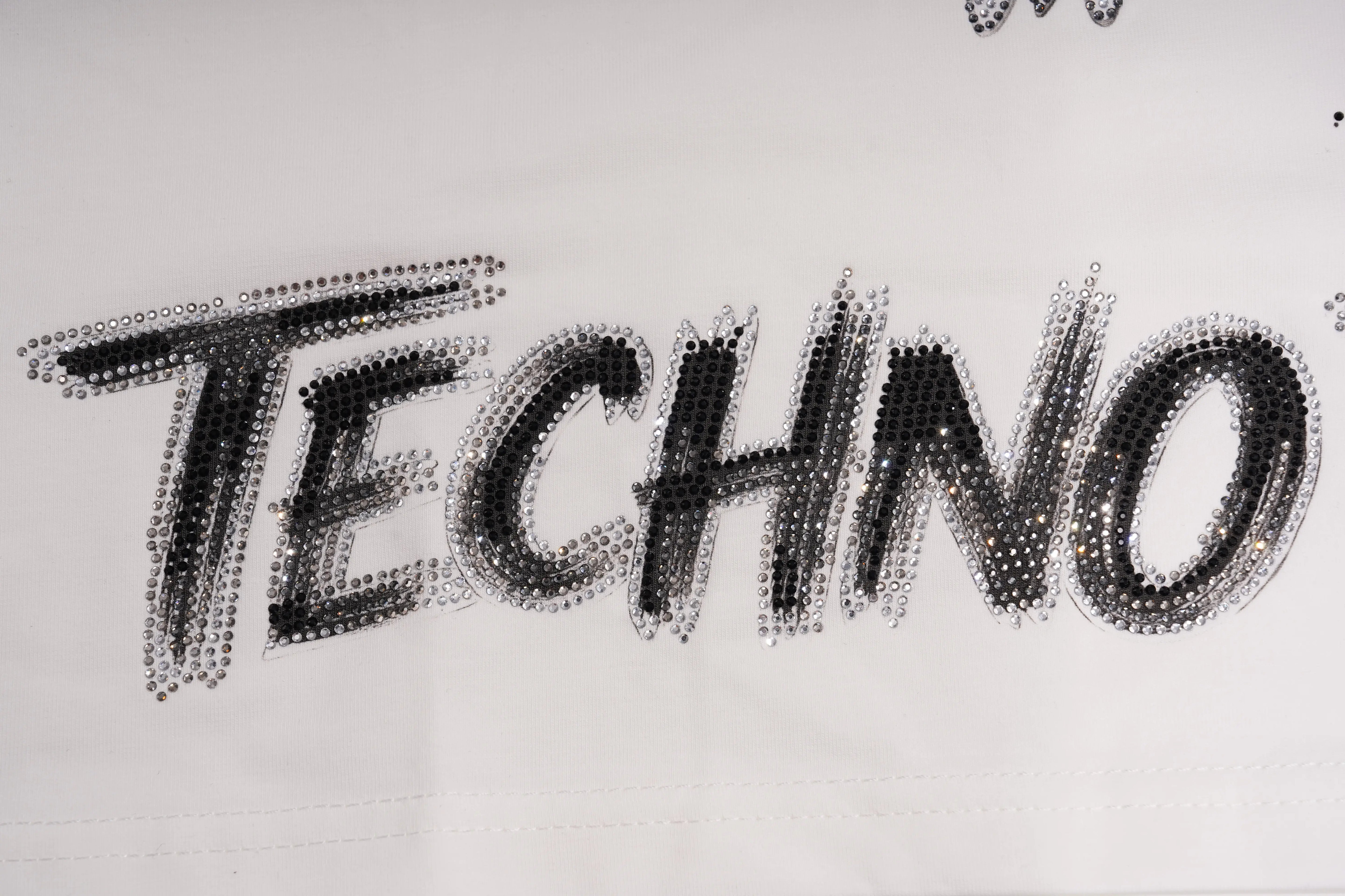 A close-up of a white fabric surface with the word "TECHNO" spelled out in large black outlined lettering made of small reflective rhinestones. TCH Brush Logo Graphic Oversized T-Shirt