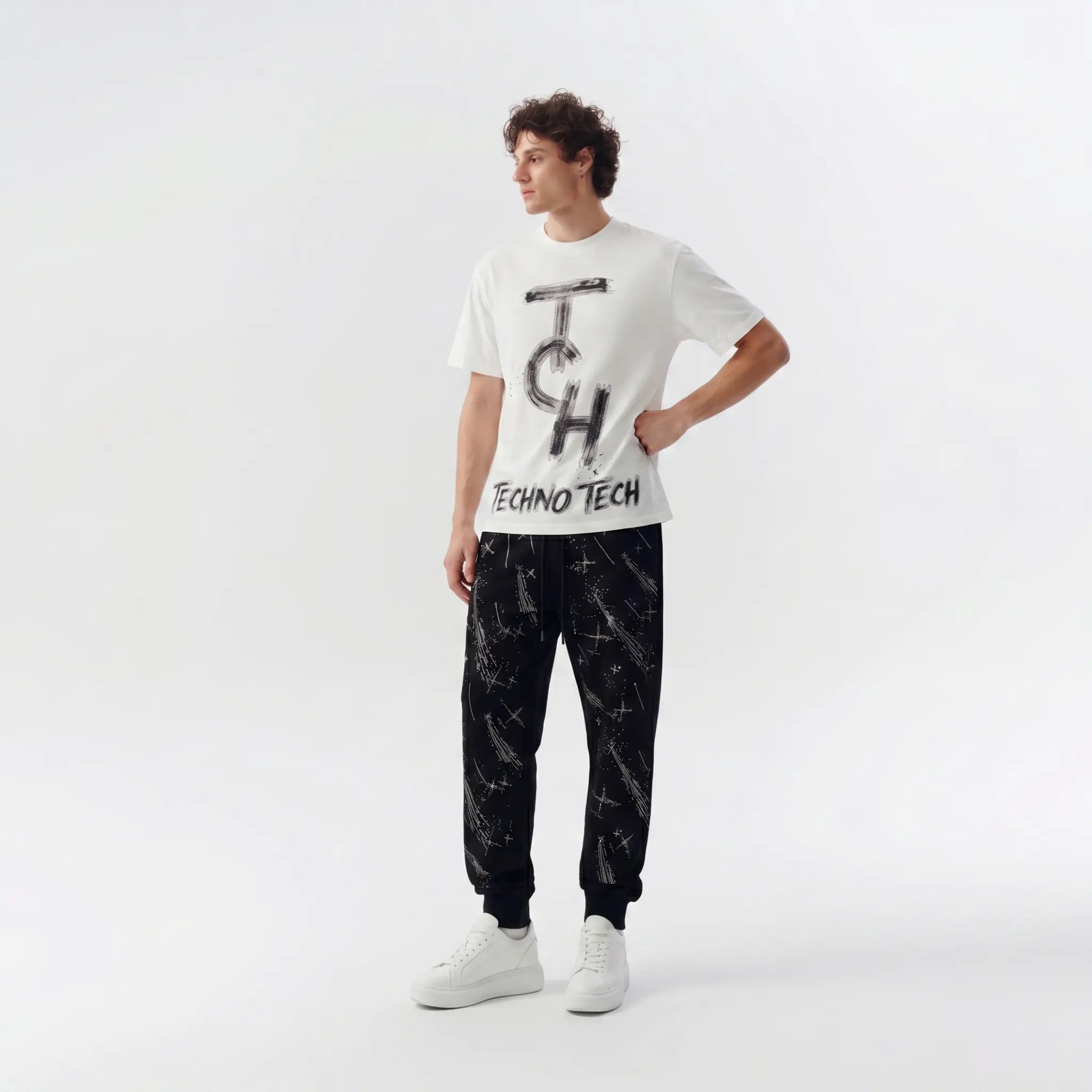 A male model stands in a studio wearing a white short-sleeve graphic T-shirt with a large black 'TCH' and 'TECHNO TECH' print, paired with black jogger pants featuring a white abstract pattern and white sneakers. TCH Brush Logo Graphic Oversized T-Shirt