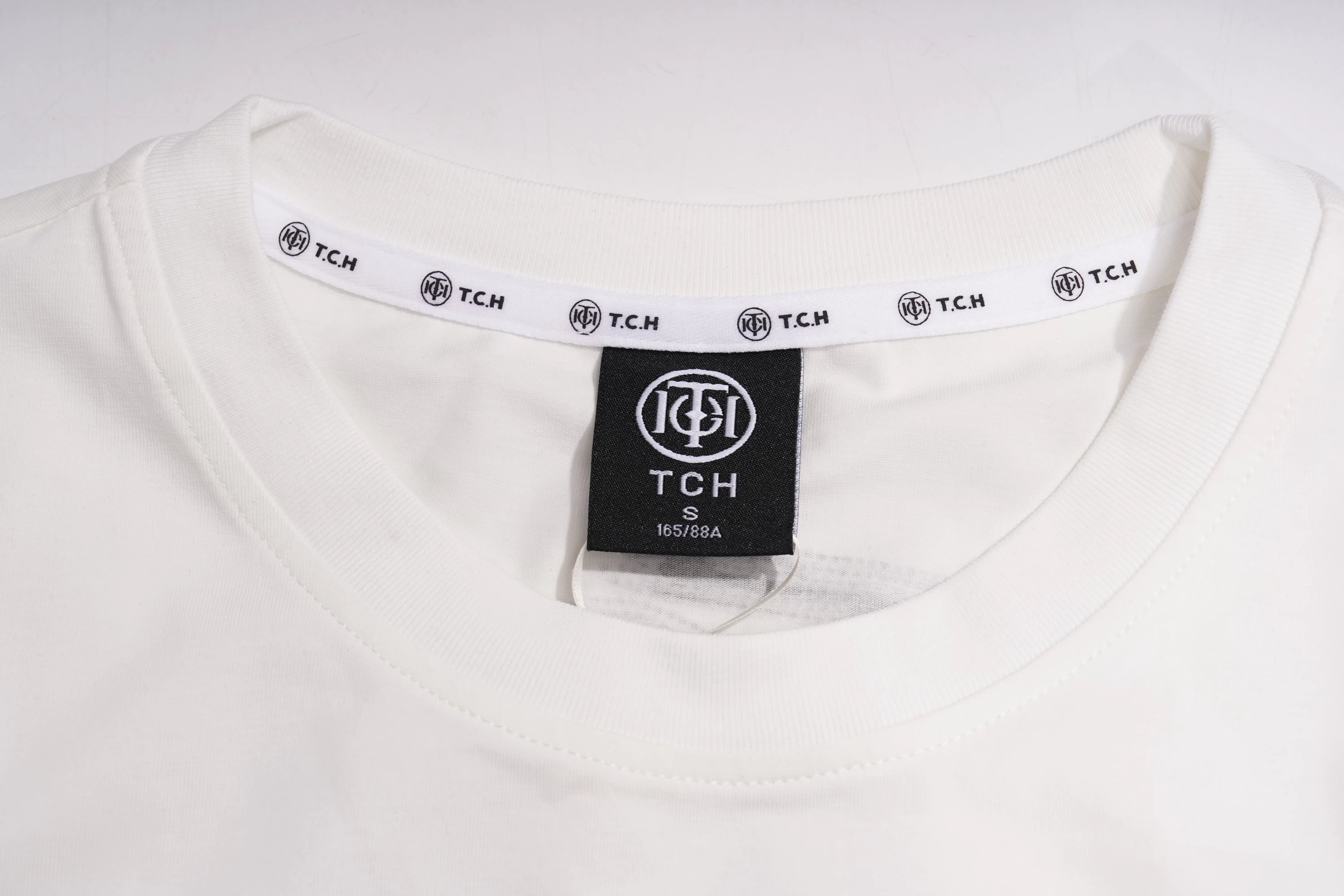 Close-up of a white crewneck t-shirt showing the inner neck label with a black TCH logo and a white inner collar band with repeating TCH logos. TCH Brush Logo Graphic Oversized T-Shirt