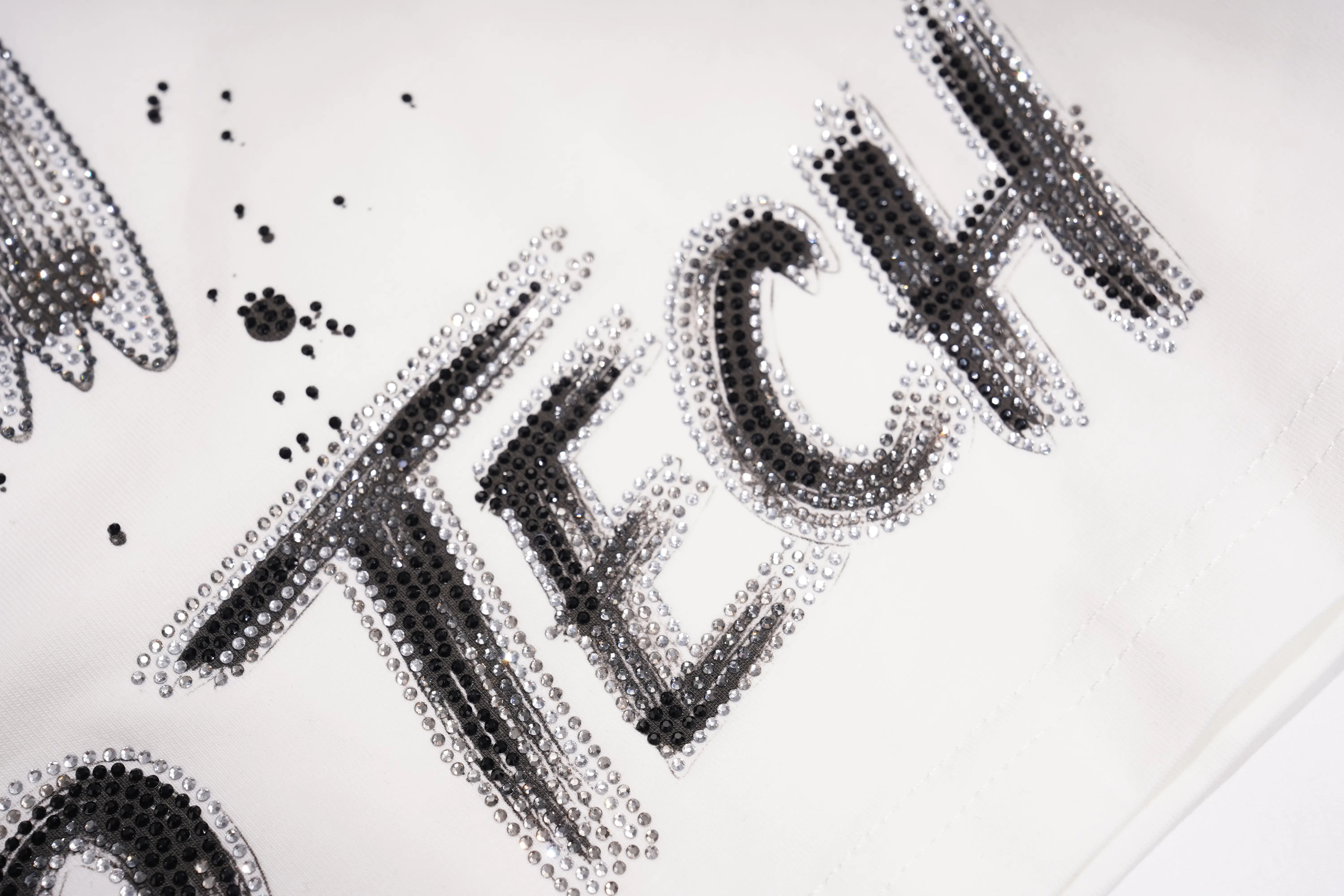 A close-up of a white surface decorated with black and silver rhinestone lettering that spells "TECH". TCH Brush Logo Graphic Oversized T-Shirt