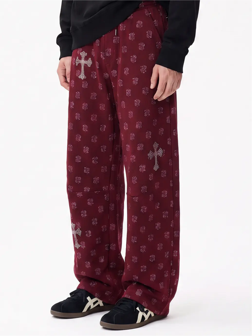 A person wearing loose burgundy pants with a repeating silver cross and floral pattern, paired with a black top and black sneakers with white logo details, shown against a plain white studio background. TCH Burgundy Paisley Print Wide-Leg Sweatpants with Cross Graphic