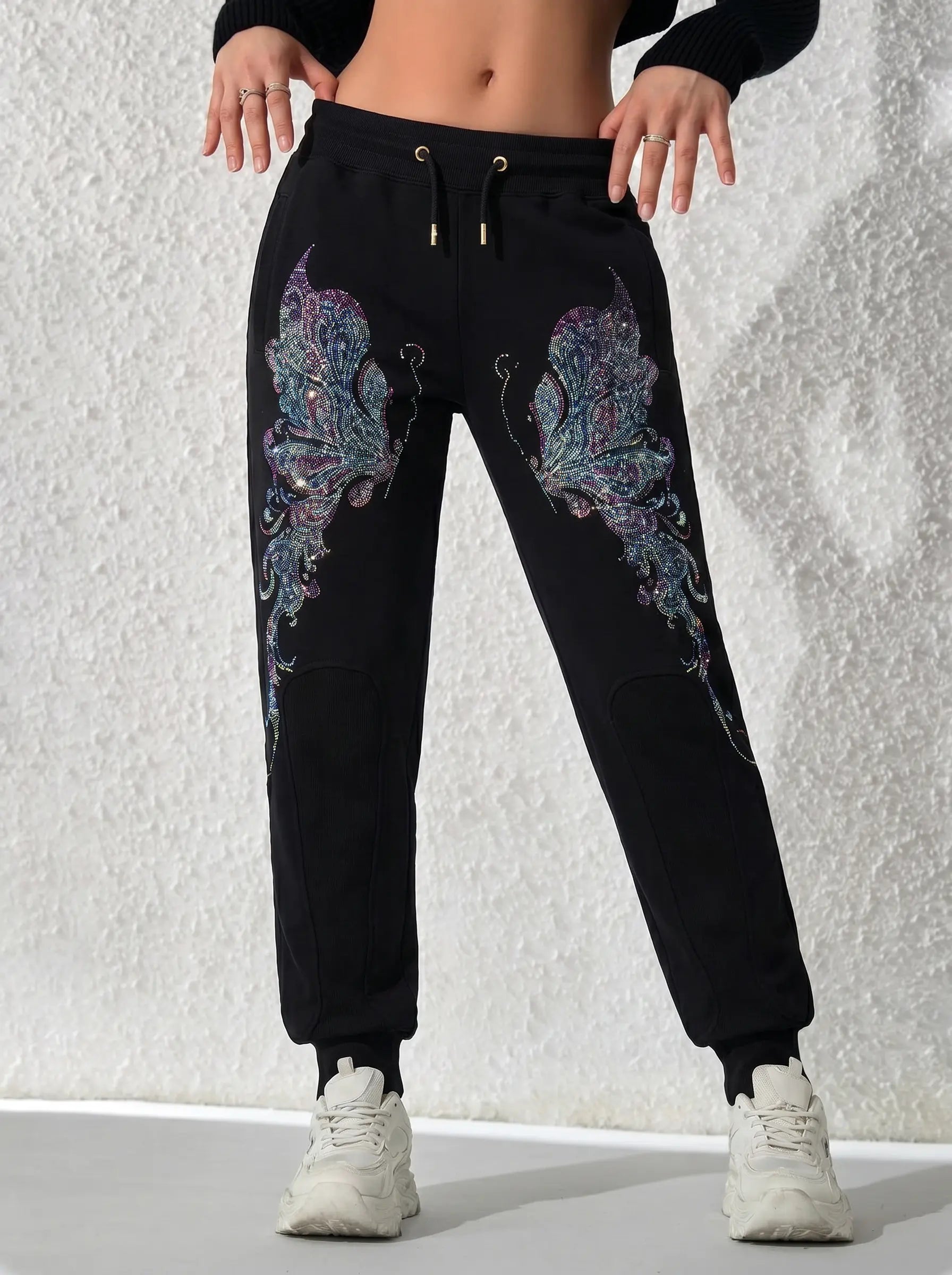 Black jogger sweatpants with large iridescent butterfly wing graphics on the thighs, shown on a model from the waist down wearing white sneakers against a textured white wall. TCH Butterfly Rhinestone Graphic Black Jogger Pants