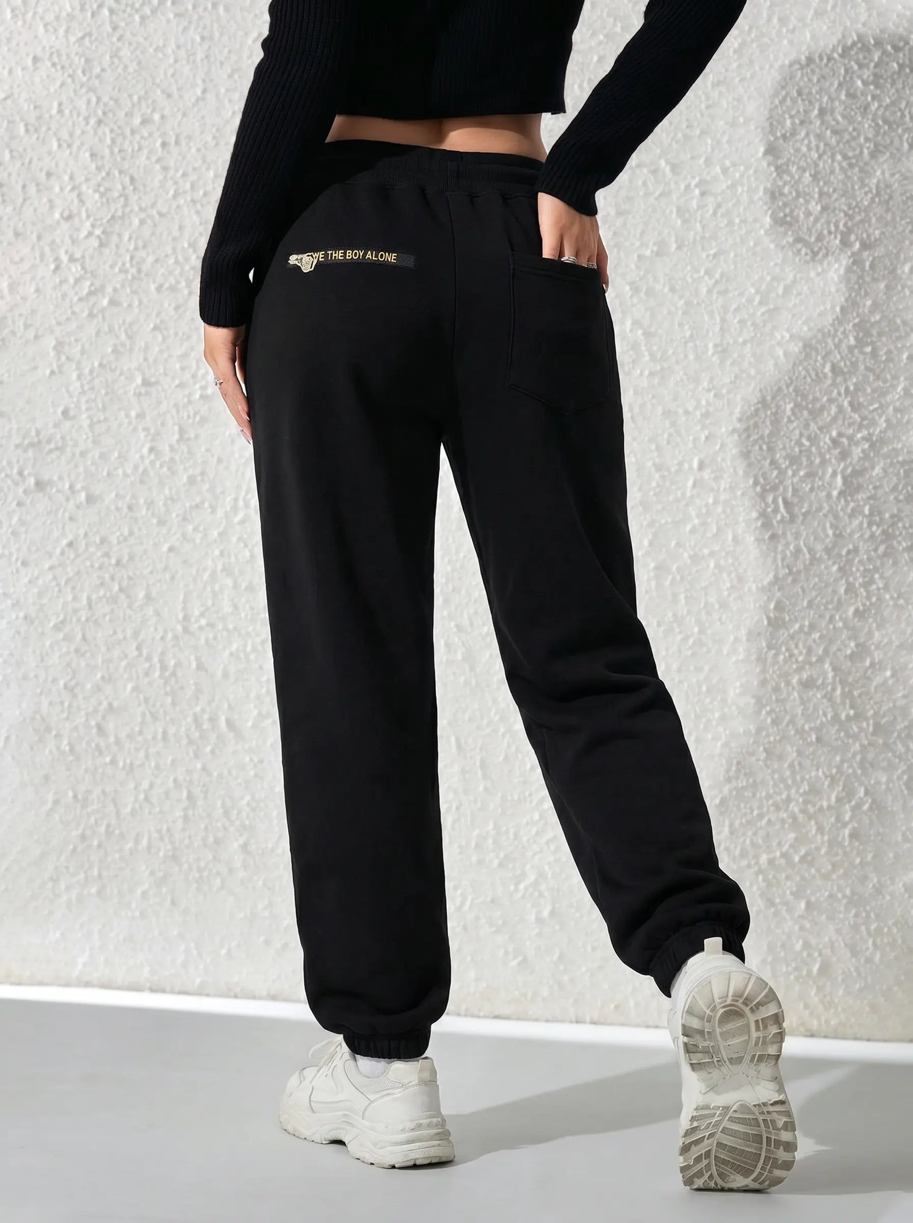 Black jogger sweatpants with a small gold embroidered logo on the back pocket, modeled from behind against a textured white wall. TCH Butterfly Rhinestone Graphic Black Jogger Pants