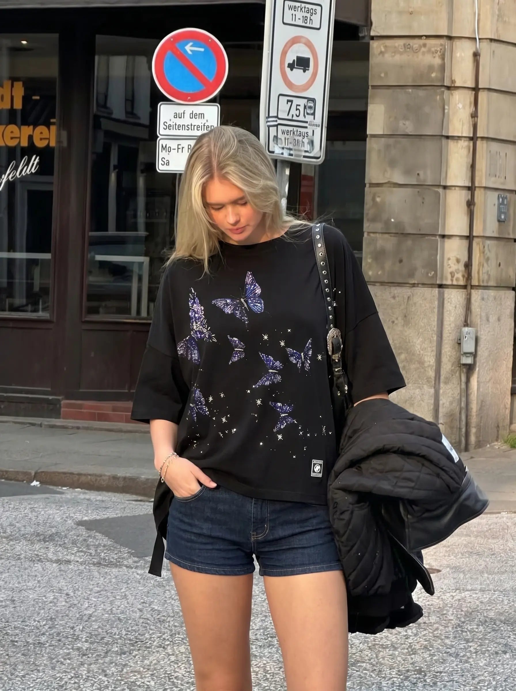 Person wearing a black t-shirt with butterfly designs and denim shorts on a street. TCH Butterfly Rhinestone Oversized T-Shirt