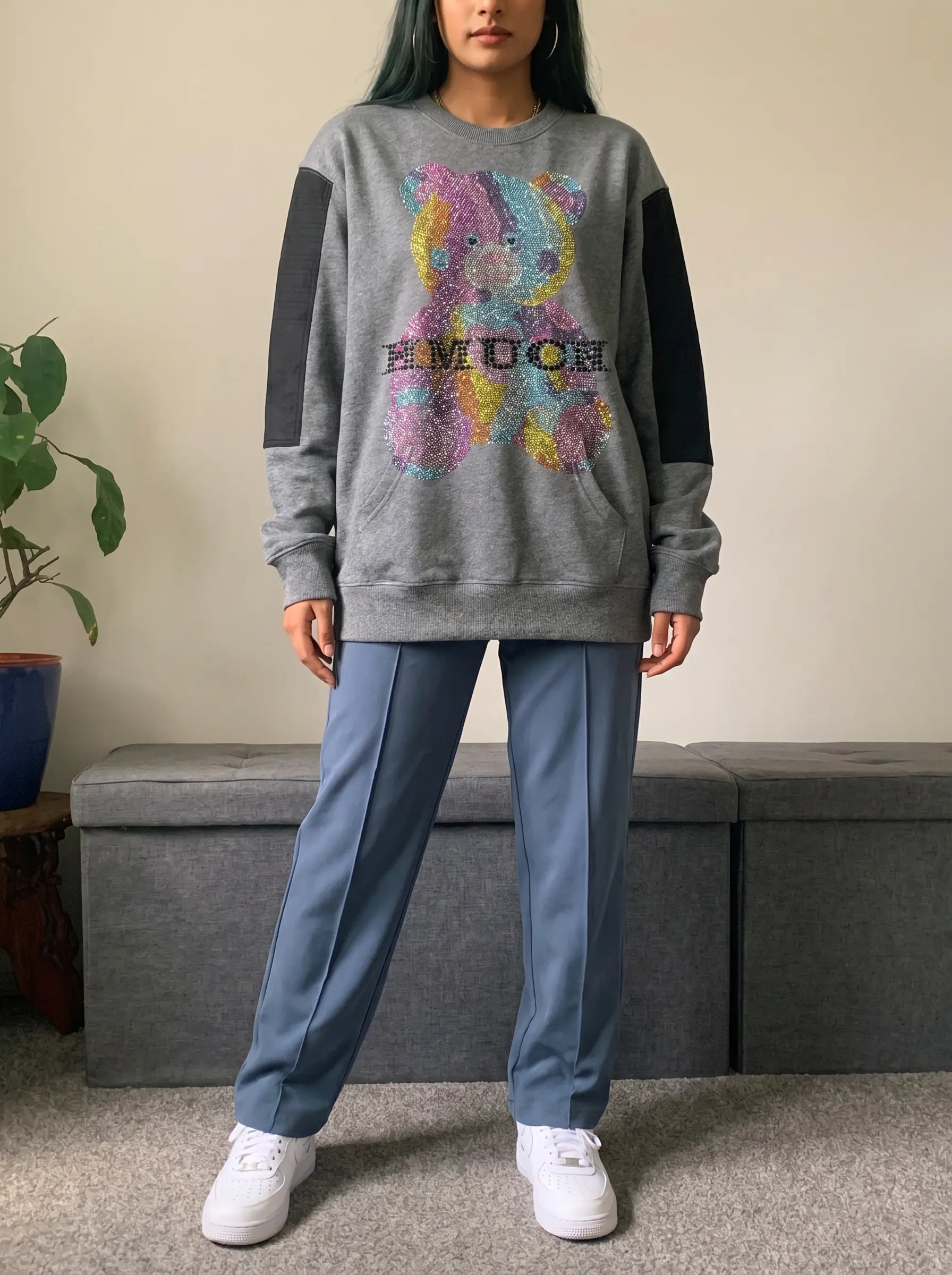 A model wearing a gray crewneck sweatshirt with black sleeve panels and a colorful sequined graphic on the front, paired with blue pants and white sneakers, standing in an indoor setting. TCH Colorful Rhinestone Teddy Bear Crewneck Sweatshirt