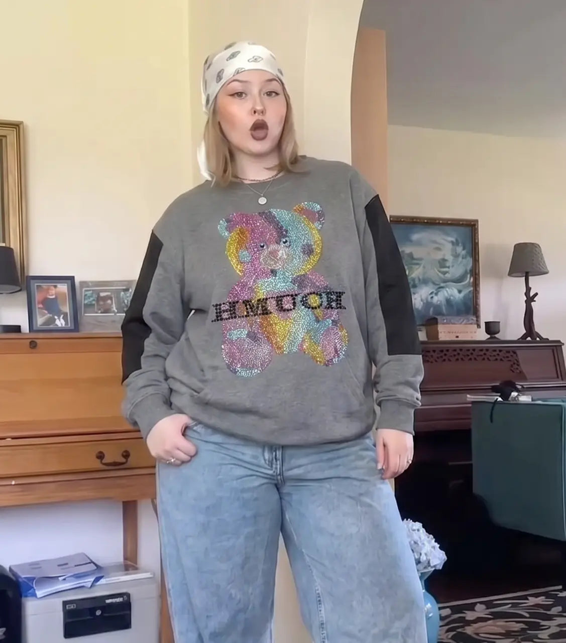 A woman wearing a gray crewneck sweatshirt with black sleeves and a colorful sequined teddy bear graphic that spells 'HMUCH', paired with light blue jeans and a white patterned headband, standing in a room with a wooden piano in the background. TCH Colorful Rhinestone Teddy Bear Crewneck Sweatshirt