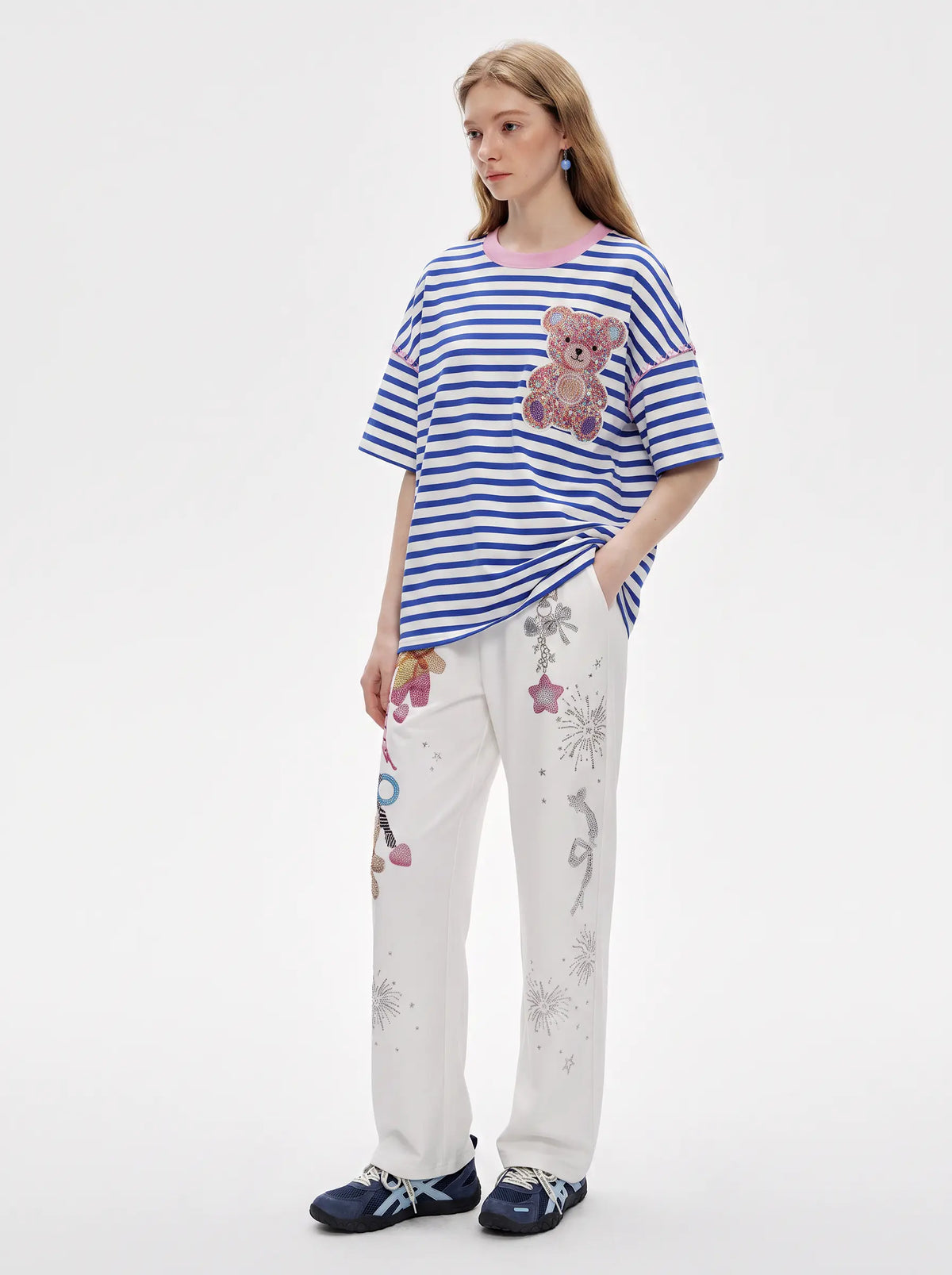 A model stands against a plain white studio background wearing a blue and white striped short-sleeve T-shirt with a teddy bear patch, white pants with floral and bird prints, and navy loafers. TCH Crystal Bear Graphic Wide-Leg Sweatpants