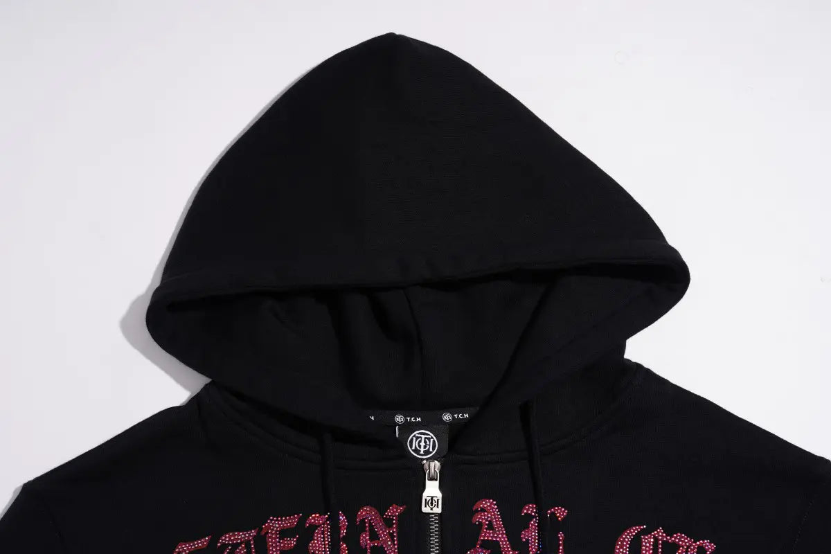A close-up of a black zip-up hoodie with a silver zipper and a red gothic-style graphic text across the chest, set against a plain white background. TCH Crystal Eagle Graphic Zip Up Hoodie