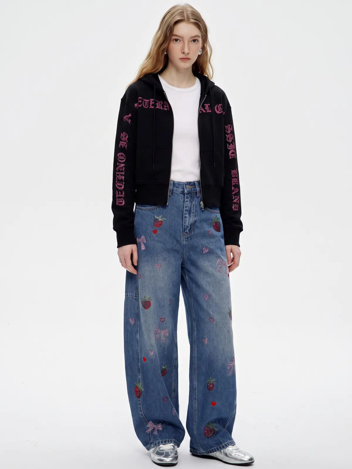 A model stands against a plain white studio background wearing a black zip-up hoodie with pink embroidered text down the sleeves, a white t-shirt, wide-leg blue jeans with strawberry and bow embroidery, and silver sneakers. TCH Crystal Eagle Graphic Zip Up Hoodie