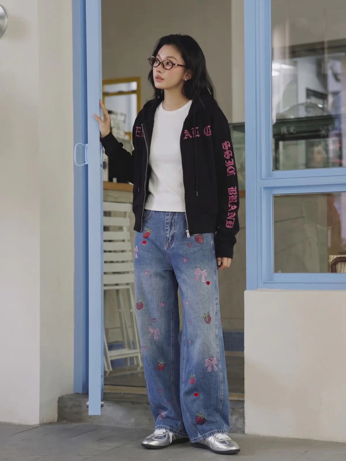 A woman wearing a black cropped zip-up jacket with pink embroidered lettering, a white ribbed top, and light blue wide-leg jeans with strawberry and bow embroidery, standing in a doorway. TCH Crystal Eagle Graphic Zip Up Hoodie