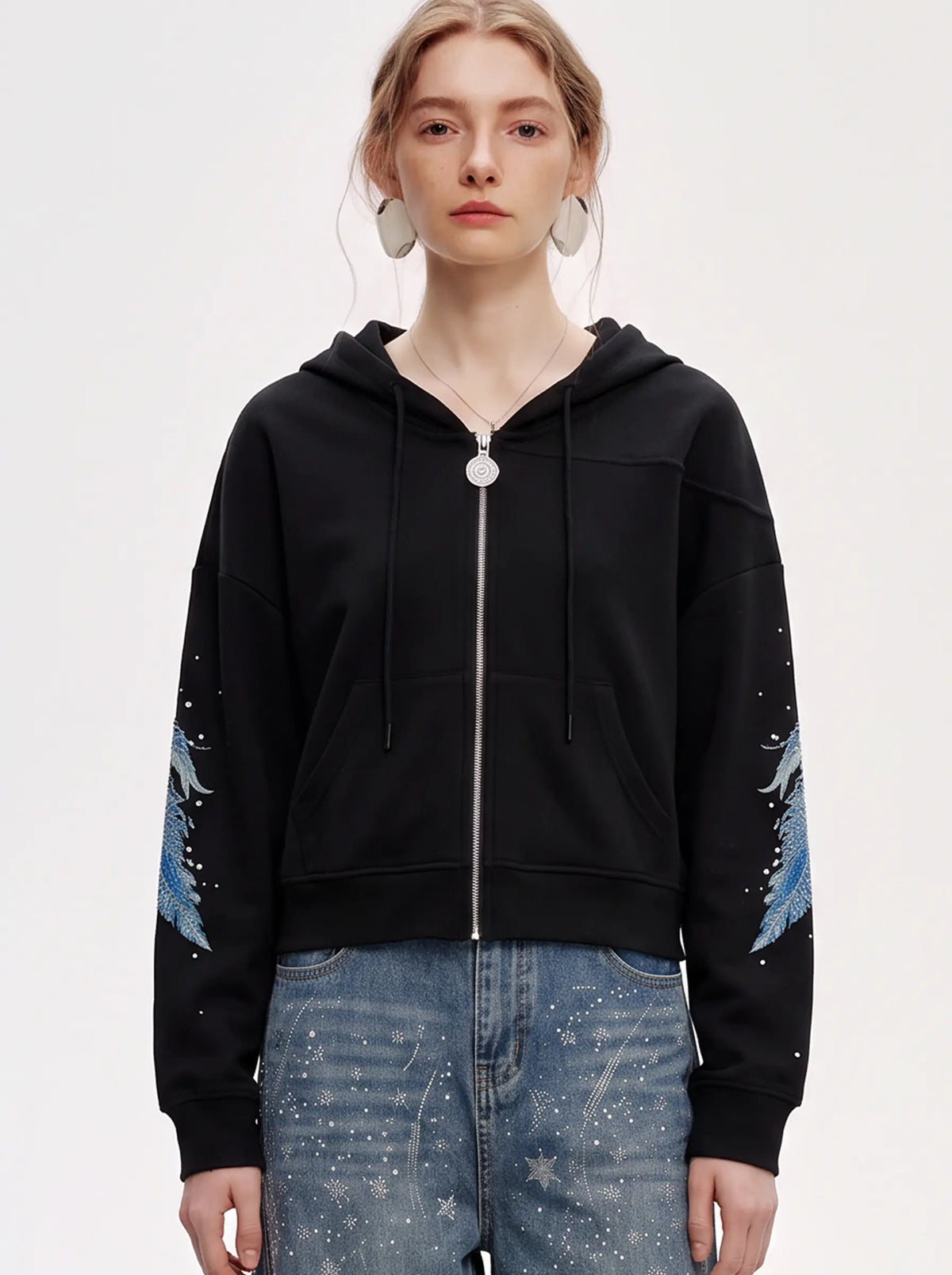 A young man wearing glasses, a light blue and white striped button-down shirt, a blue patterned necktie, and gray jeans, standing against a plain off-white studio background. TCH Crystal Wing Graphic Zip Up Hoodie