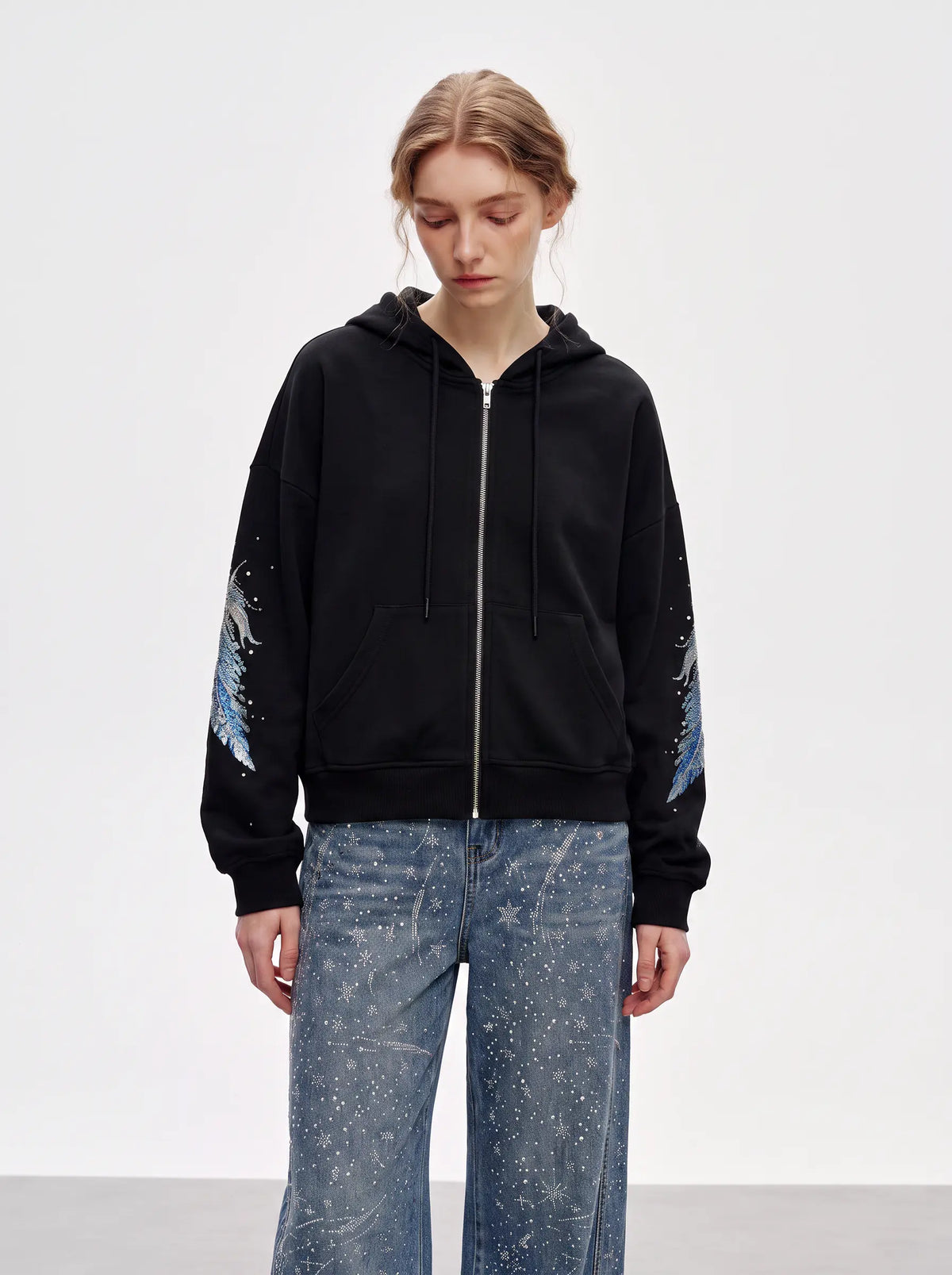 A model wearing a black zip-up hoodie with blue and white graphic prints on the sleeves, paired with blue paint-splattered jeans, against a plain white studio background. TCH Crystal Wing Graphic Zip Up Hoodie