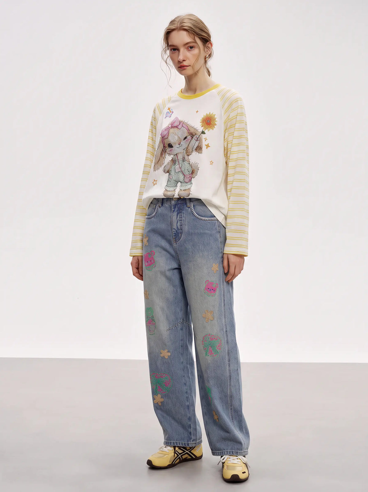 A model stands in a studio wearing a cream and yellow striped long-sleeve sweater with a bunny graphic, light blue wide-leg jeans with colorful embroidered details, and yellow sneakers. TCH Cute Bunny Rhinestone Graphic Striped Sleeve Long Sleeve T-Shirt
