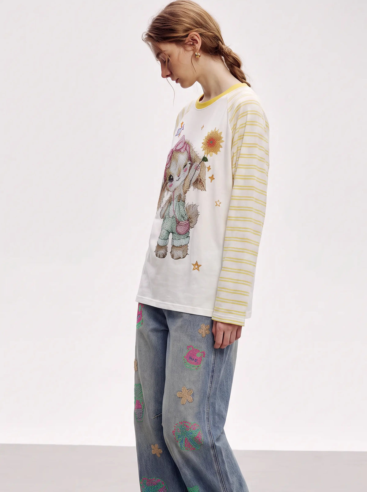 A model wearing a long-sleeve white t-shirt with a cartoon bunny graphic, yellow striped sleeves, and blue jeans with colorful embroidered flowers, set against a plain light gray studio background. TCH Cute Bunny Rhinestone Graphic Striped Sleeve Long Sleeve T-Shirt