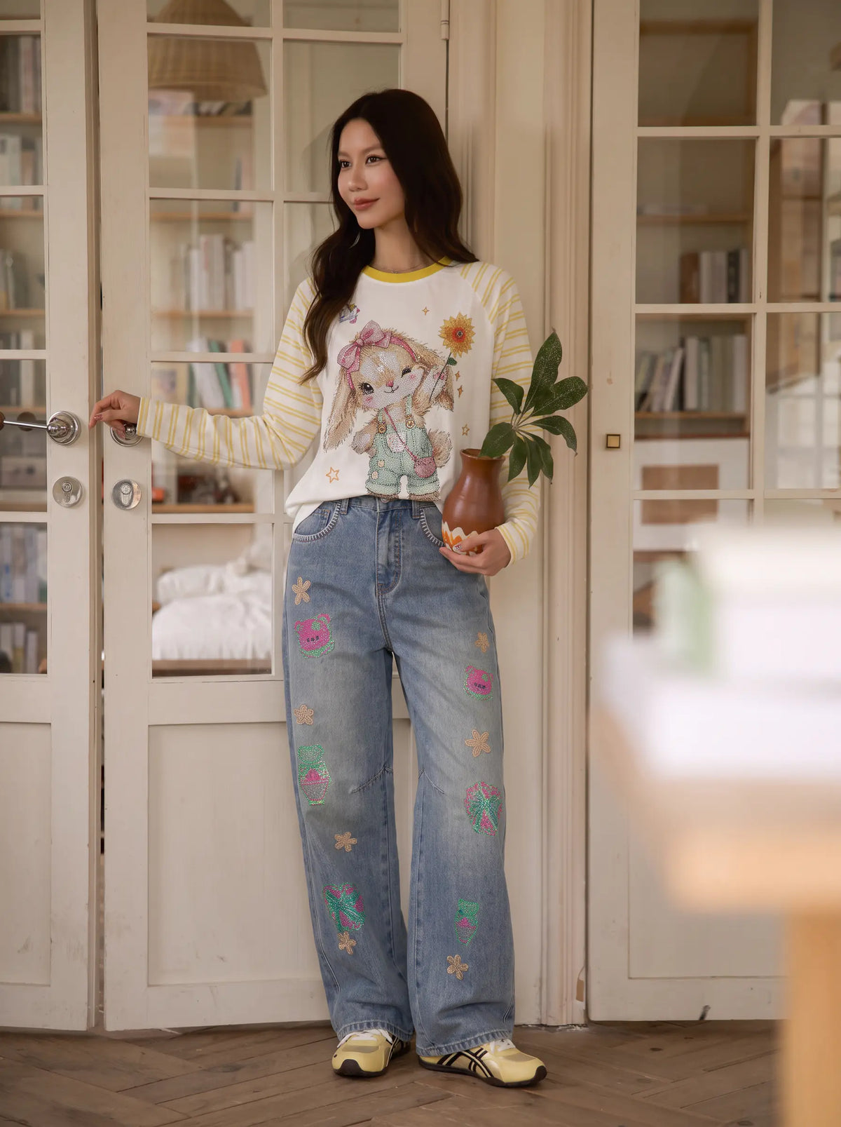 A woman stands in front of cream-colored glass-paneled doors, wearing a white and yellow striped long-sleeve shirt with a cartoon girl graphic, light-wash blue jeans with colorful embroidered flowers, and yellow sneakers. She holds a small brown vase with green leaves. TCH Cute Bunny Rhinestone Graphic Striped Sleeve Long Sleeve T-Shirt