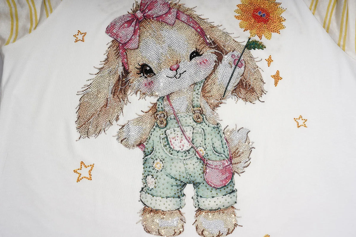 A close-up of a white fabric item, likely a pillowcase or blanket, featuring a detailed cross-stitch style illustration of a cute beige bunny wearing green polka-dot overalls, a pink bow, and holding a yellow flower. TCH Cute Bunny Rhinestone Graphic Striped Sleeve Long Sleeve T-Shirt
