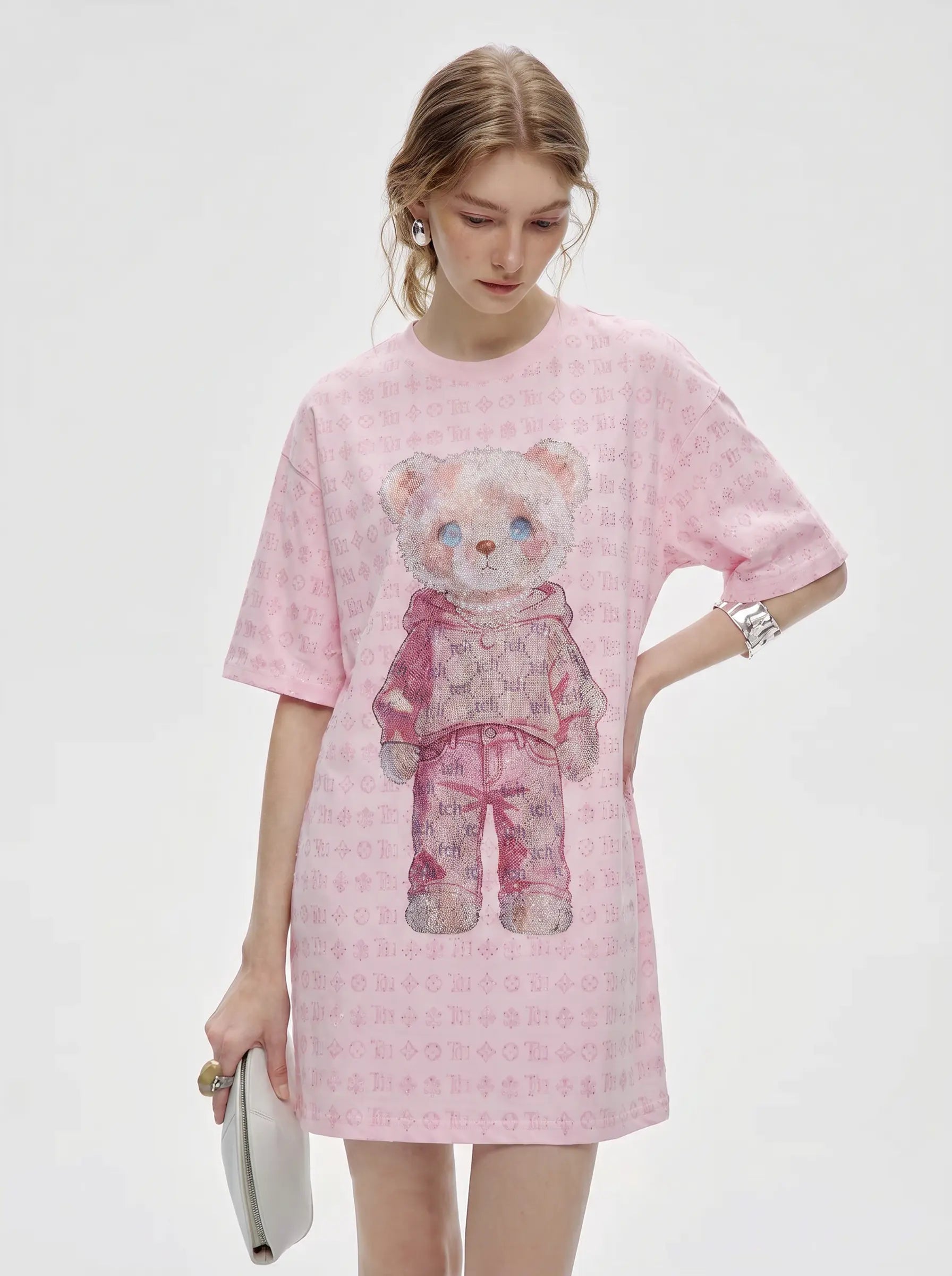 A model wearing a pink short-sleeve t-shirt dress with a repeating monogram pattern and a large teddy bear graphic on the front, holding a silver clutch against a light gray background. TCH Cute Hoodie Teddy Bear Rhinestone Oversized T-Shirt Dress