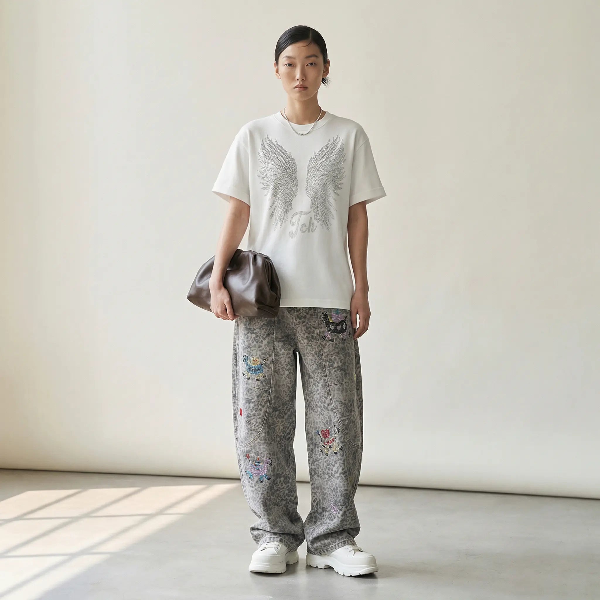 Person wearing a white t-shirt with a graphic design and patterned pants, holding a brown leather bag. TCH Cute Pony Rhinestone Grey Leopard Wide-Leg Jeans