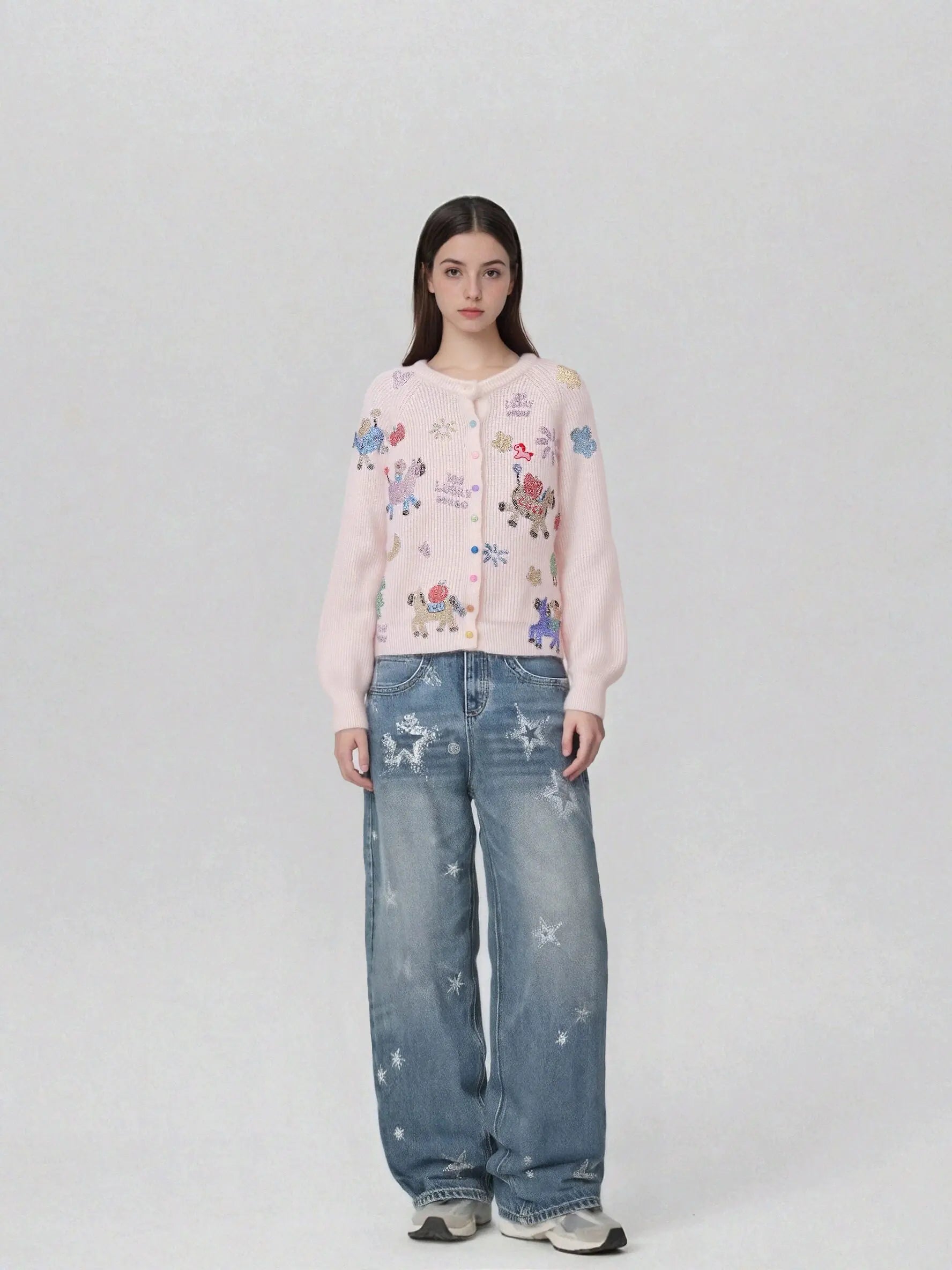 A model stands against a light gray studio background wearing a light pink cropped hoodie with colorful cartoon unicorn prints and distressed blue wide-leg jeans. TCH Cute Rhinestone Animal Garden Knit Cardigan