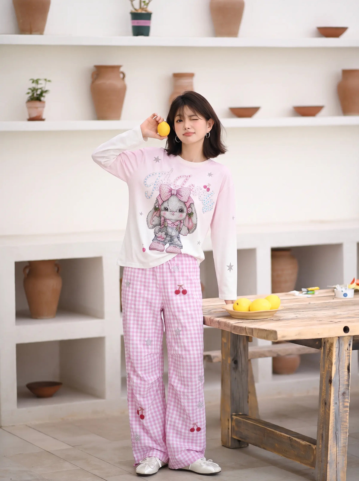 A woman stands in a bright room wearing a two-piece pajama set consisting of a light pink long-sleeve top with a cartoon bunny graphic and matching pink gingham pants with cherry prints. She is holding a yellow fruit near her face. TCH Cute Rhinestone Cat Graphic Pink Long Sleeve T-Shirt