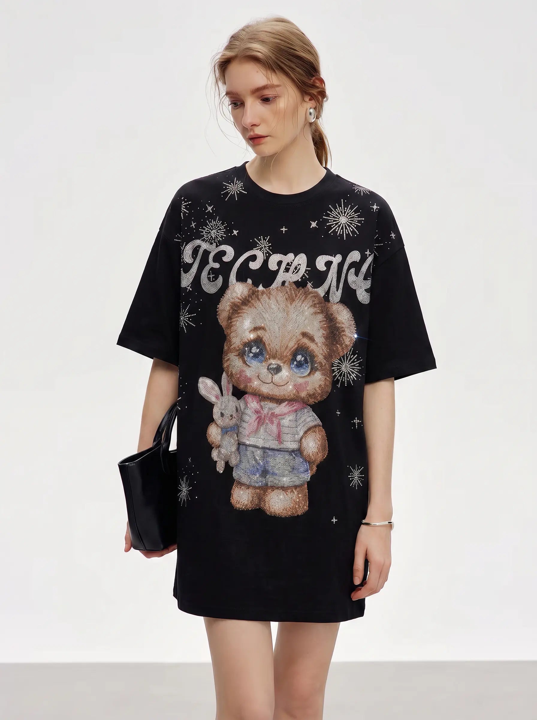 A woman modeling a black oversized graphic T-shirt dress featuring a teddy bear illustration, white decorative text, and white snowflake motifs, against a plain light gray studio background. TCH Cute Teddy Bear Rhinestone Oversized T-Shirt Dress