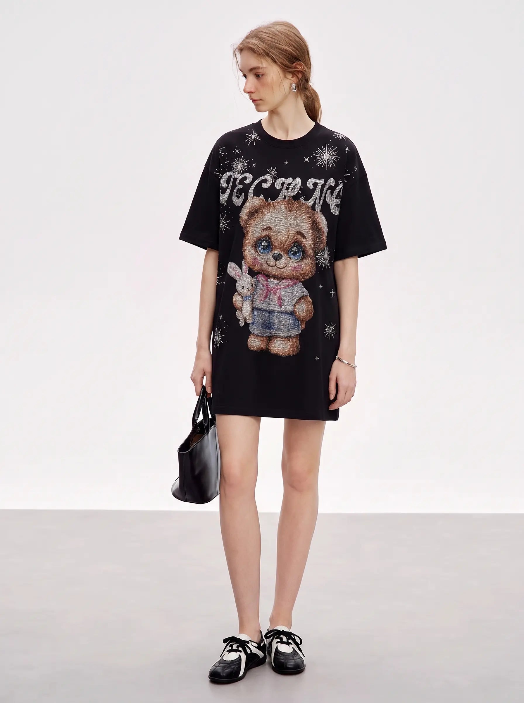 A model stands against a light gray studio background wearing an oversized black graphic T-shirt dress with a teddy bear print and white text, holding a black handbag and wearing black-and-white sneakers. TCH Cute Teddy Bear Rhinestone Oversized T-Shirt Dress