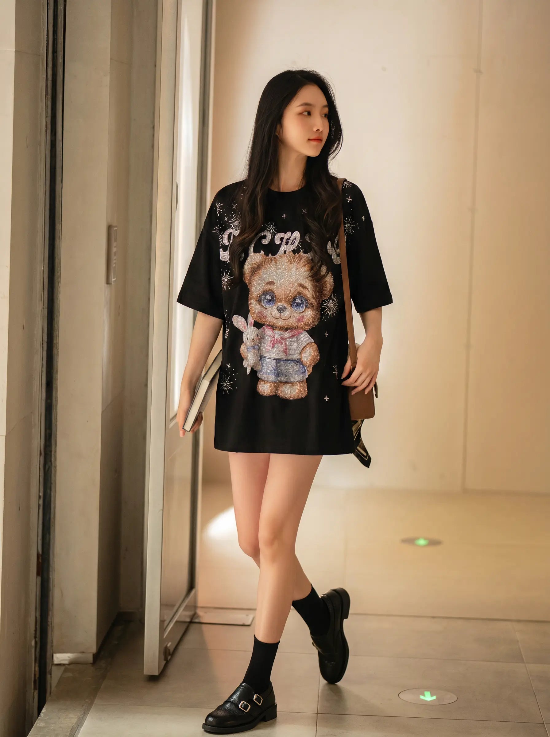 A woman wearing a black oversized graphic T-shirt dress with a large cartoon character print on the front, styled with black socks and black loafers, walking in a modern indoor hallway. TCH Cute Teddy Bear Rhinestone Oversized T-Shirt Dress