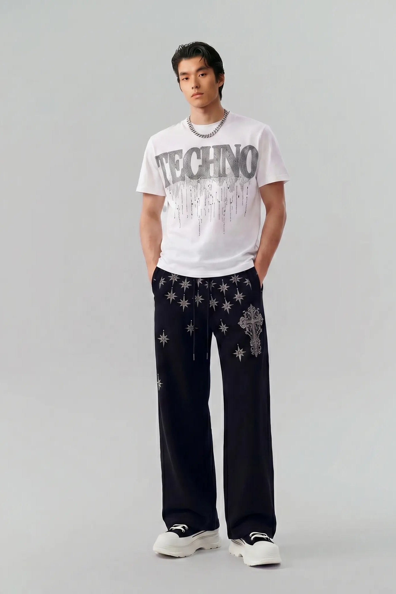 A male model stands against a light gray studio background wearing a white graphic T-shirt with 'TECHNO' text, loose black pants with silver star and cross embellishments, and white sneakers. TCH Dripping Rhinestone Logo Graphic T-Shirt