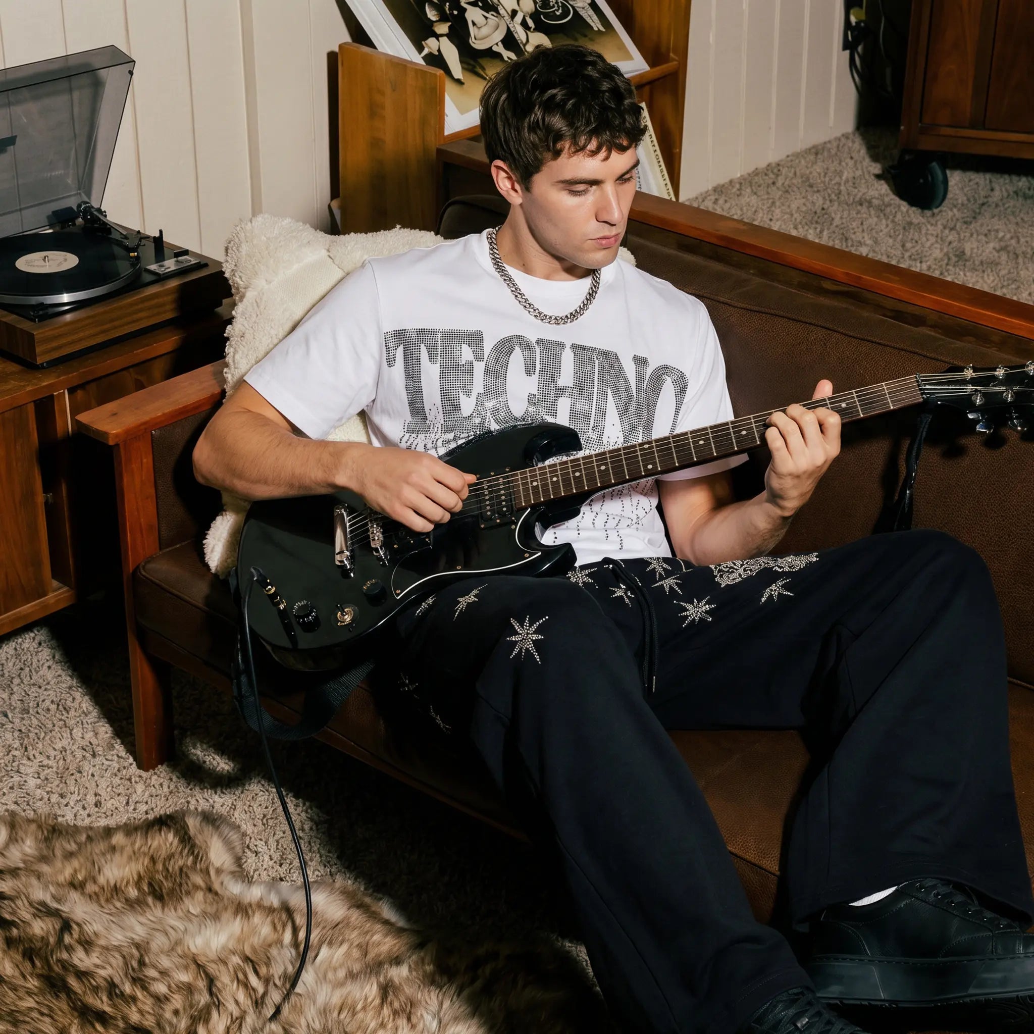A man sits on a brown leather couch in a cozy room, playing a black electric guitar. He is wearing a white graphic t-shirt with 'TECHNO' printed on the front, dark pants with white star-like patterns, and a silver chain necklace. TCH Dripping Rhinestone Logo Graphic T-Shirt