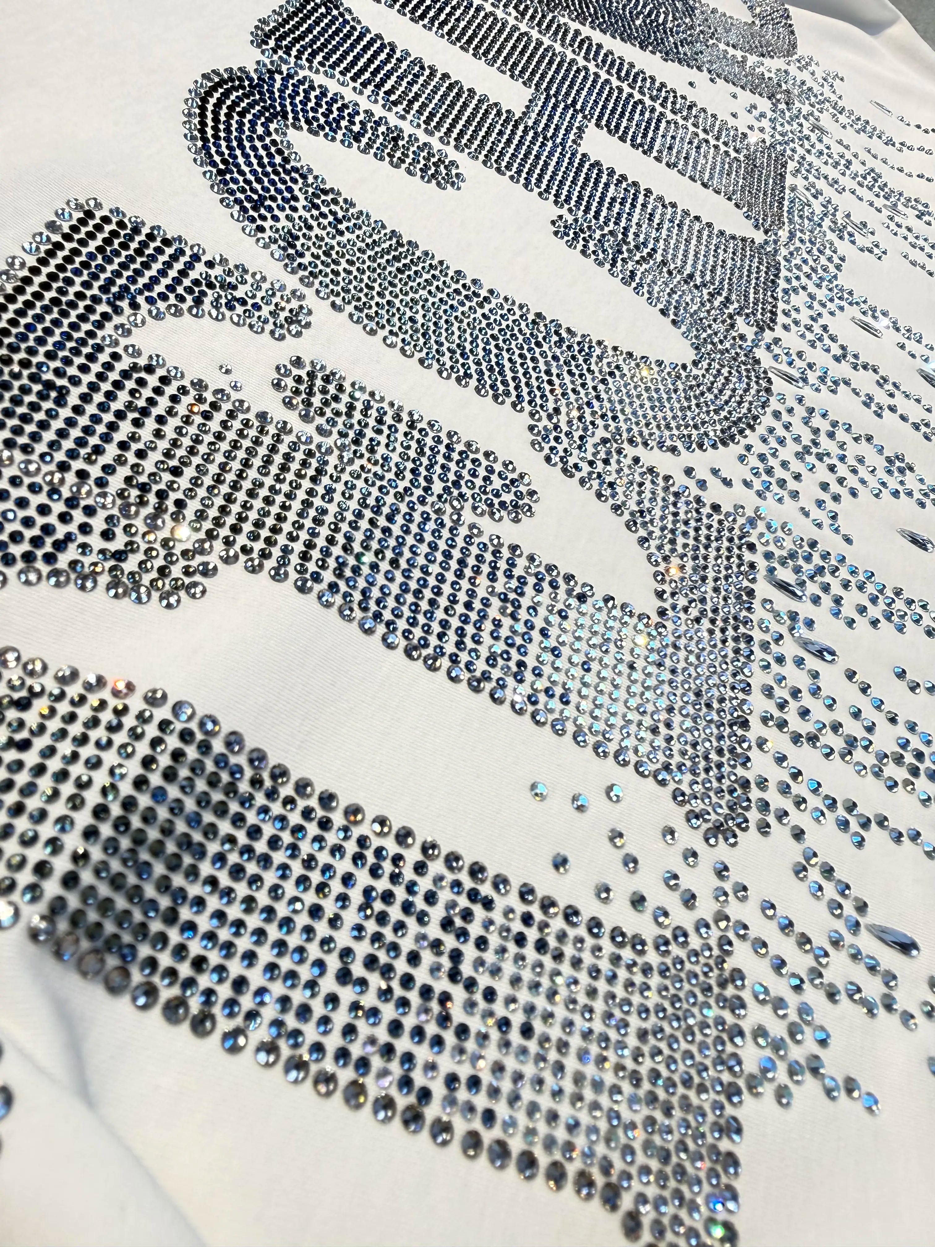 A close-up of a white garment covered with large silver rhinestone lettering, showing the texture and sparkle of the crystals. TCH Dripping Rhinestone Logo Graphic T-Shirt