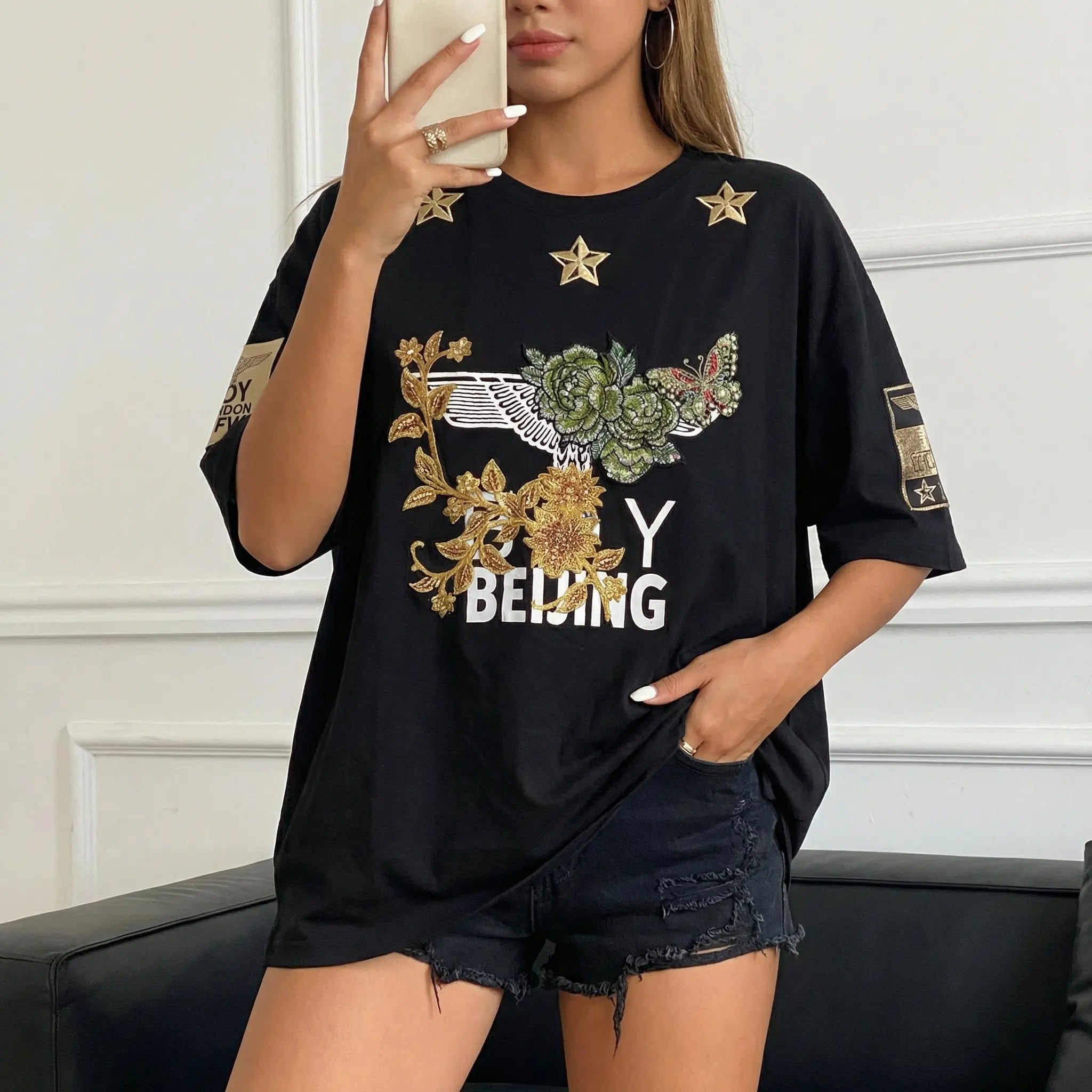 Person wearing a black t-shirt with floral and text design, sitting on a couch. TCH Embroidered Floral Butterfly Rhinestone T-Shirt