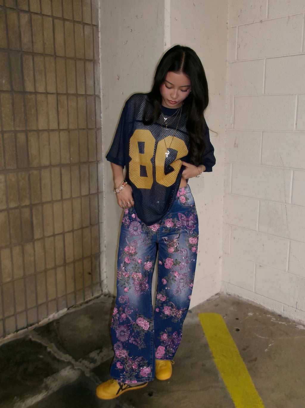 A woman stands in a concrete corner wearing a navy blue oversized graphic t-shirt with a large yellow '86' on the front, paired with blue floral wide-leg jeans and yellow shoes. TCH Floral Butterfly Rhinestone Wide-Leg Denim Jeans