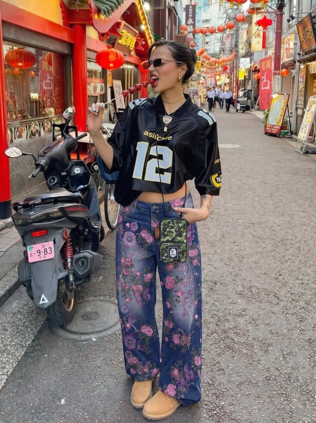 A woman posing on a street wearing a black cropped football jersey with the number 12, floral wide-leg jeans, and tan boots, with a small green camouflage pouch hanging from her waist. TCH Floral Butterfly Rhinestone Wide-Leg Denim Jeans