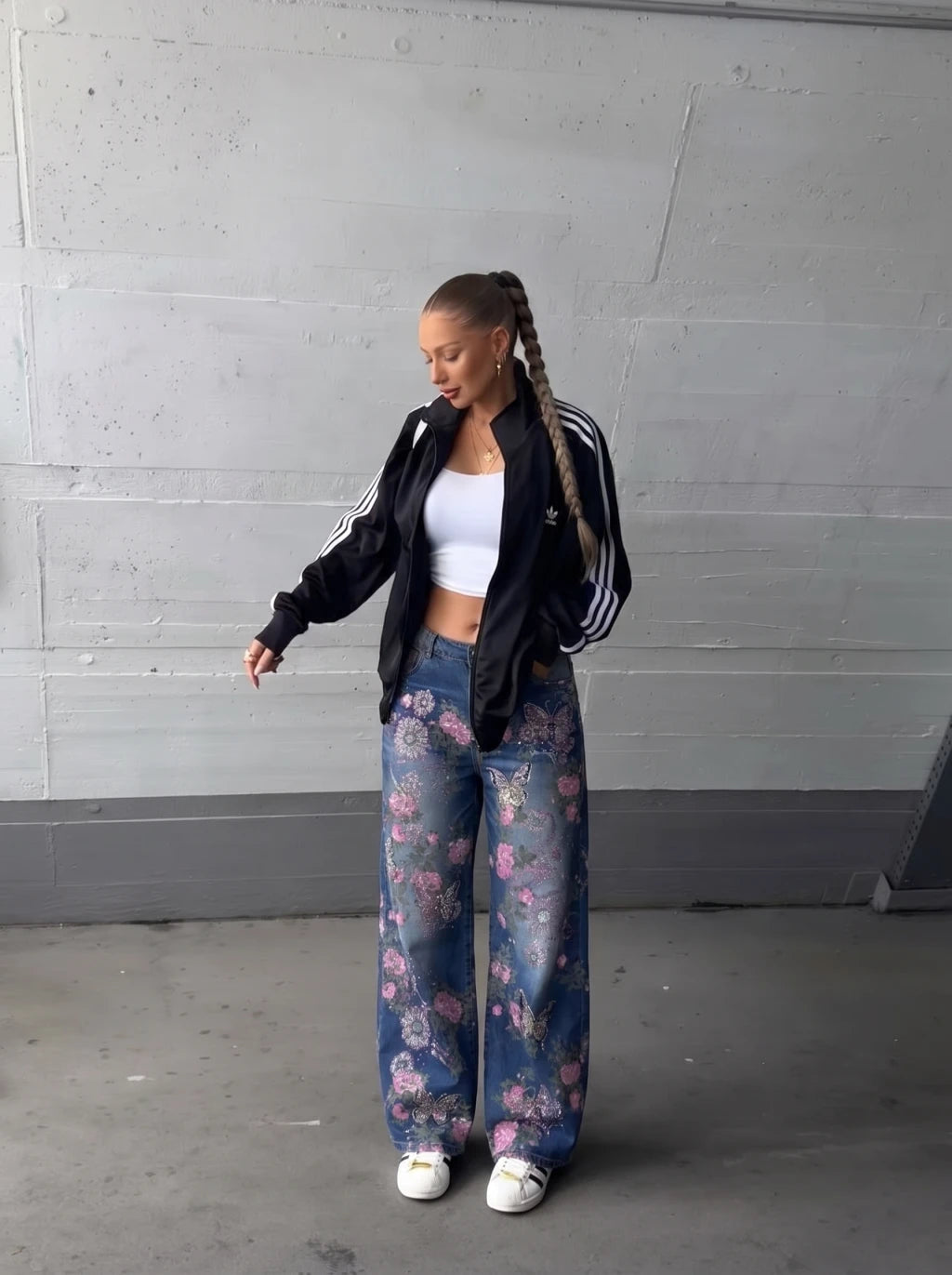 A woman stands against a gray concrete wall wearing a black Adidas track jacket with white stripes, a white crop top, wide-leg floral blue jeans, and white sneakers. TCH Floral Butterfly Rhinestone Wide-Leg Denim Jeans