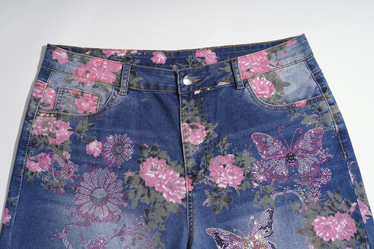 A close-up of blue denim jeans featuring a pink and purple floral print with butterfly motifs, laid flat against a light gray background. TCH Floral Butterfly Rhinestone Wide-Leg Denim Jeans