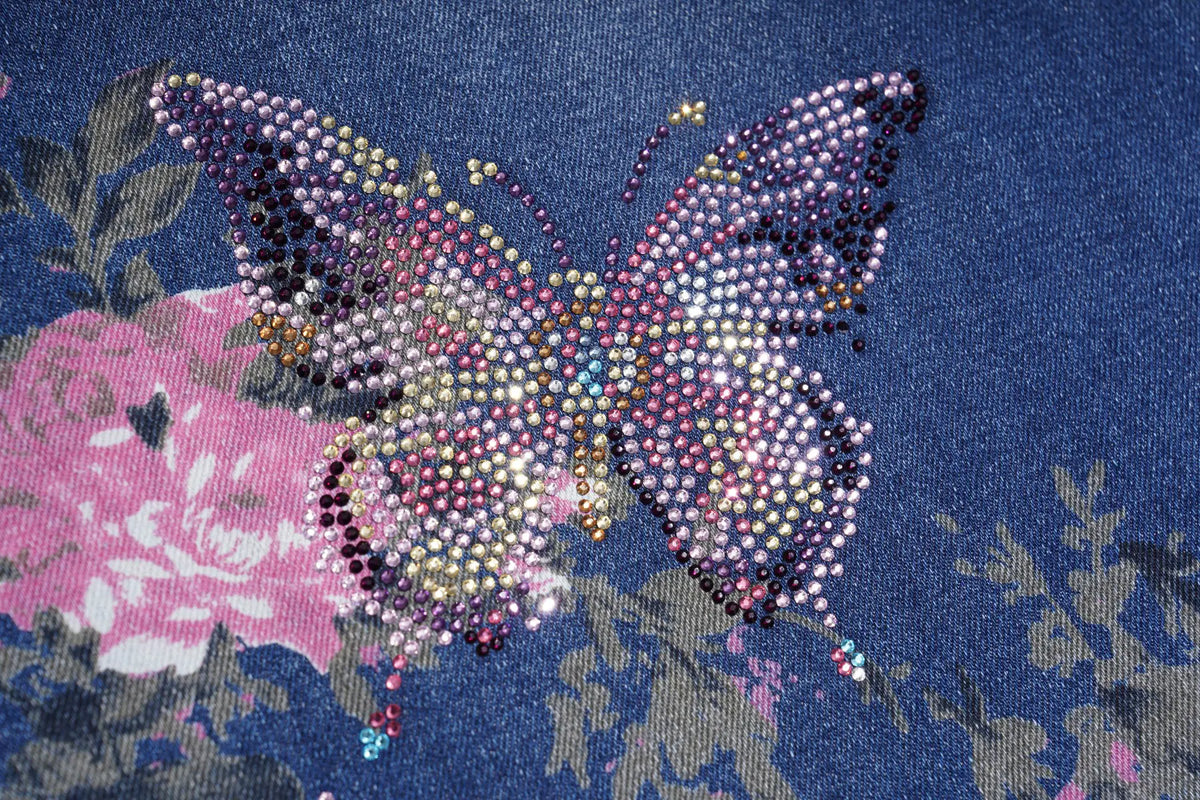 A close-up of a butterfly embellished with multicolored rhinestones on a dark blue fabric background with a pink floral pattern. TCH Floral Butterfly Rhinestone Wide-Leg Denim Jeans