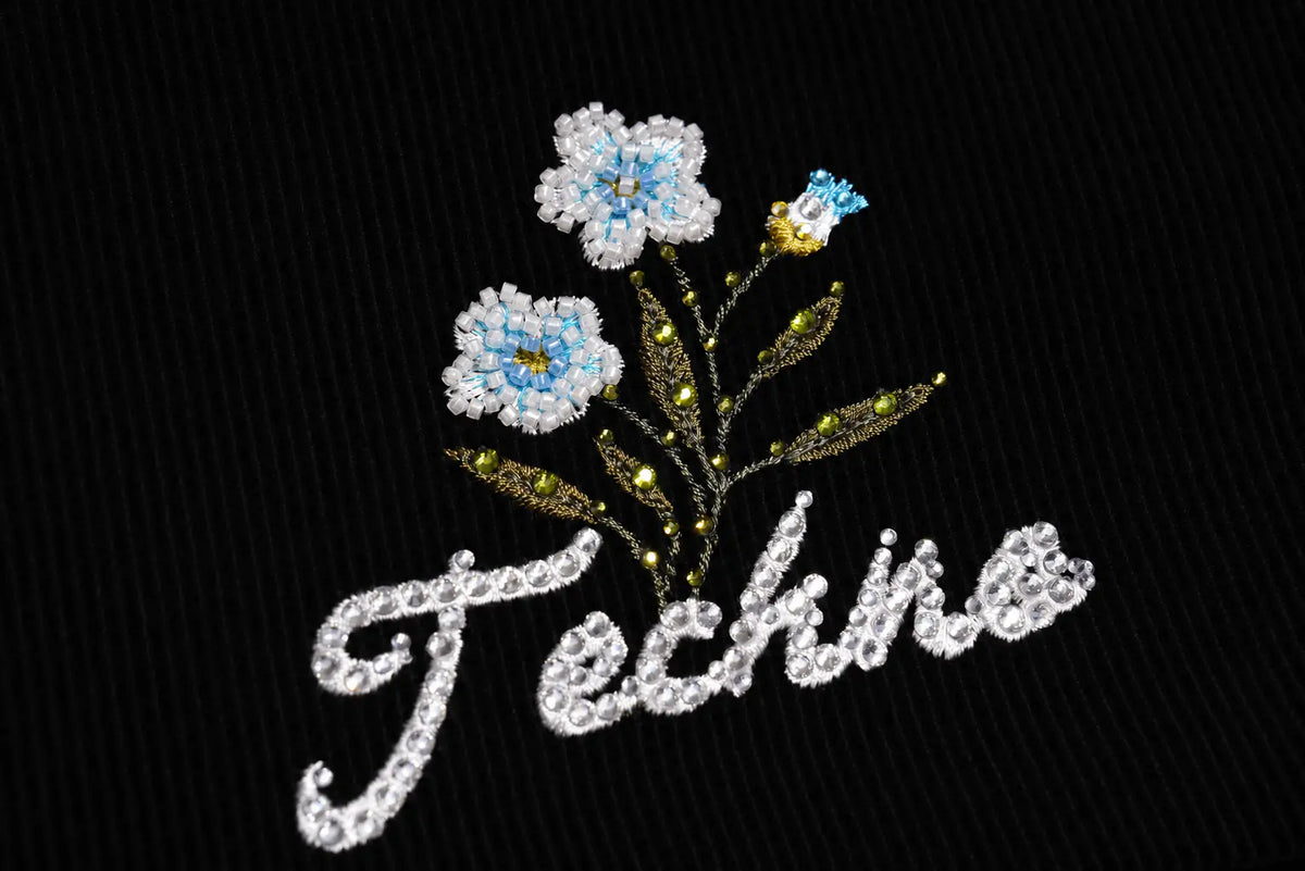 A close-up of a black ribbed fabric featuring an embroidered design with two white and light blue flowers, green and gold leaves, and the word 'Techno' in white cursive script. TCH Floral Embroidered Techno Corduroy Mini Skirt