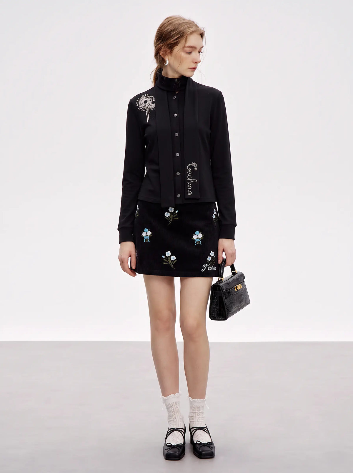 A model stands in a studio wearing a black long-sleeve button-front jacket with floral embroidery and a matching black mini skirt with embroidered floral details. She is also wearing white socks and black Mary Jane shoes, and holding a small black handbag. TCH Floral Embroidered Techno Corduroy Mini Skirt