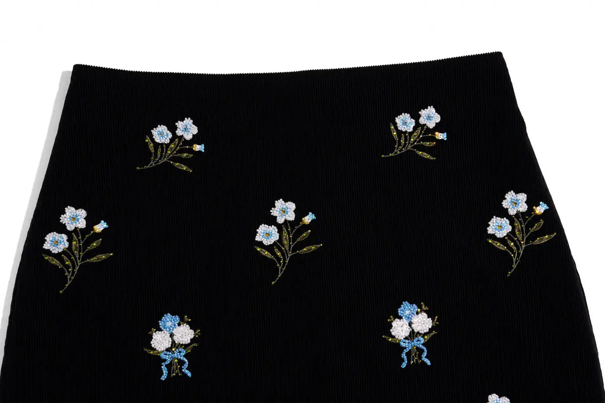 A close-up of a black ribbed knit garment, likely a skirt, featuring embroidered bouquets of light blue and white flowers with green stems and yellow accents. TCH Floral Embroidered Techno Corduroy Mini Skirt