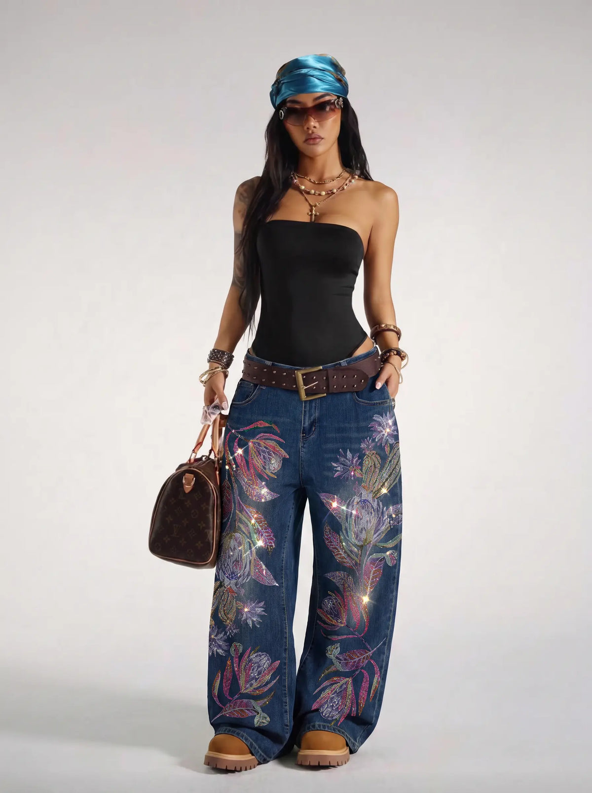 A model stands against a light gray studio background wearing a black strapless top, wide-leg blue jeans with colorful floral embroidery, a brown belt, brown boots, a blue headscarf, and holding a brown monogram handbag. TCH Floral Print Wide-Leg Denim Jeans