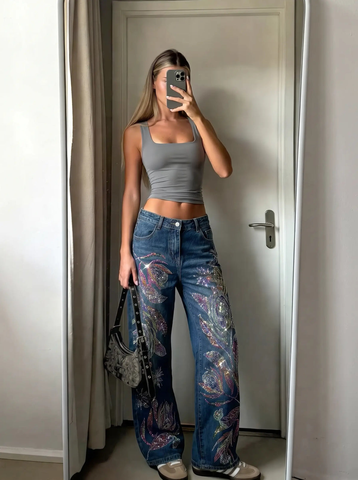 A woman takes a mirror selfie in an indoor room, wearing a gray sleeveless crop top and wide-leg blue jeans with colorful paisley patterns on the lower legs. She is holding a phone to block the mirror and carrying a small dark handbag. TCH Floral Print Wide-Leg Denim Jeans