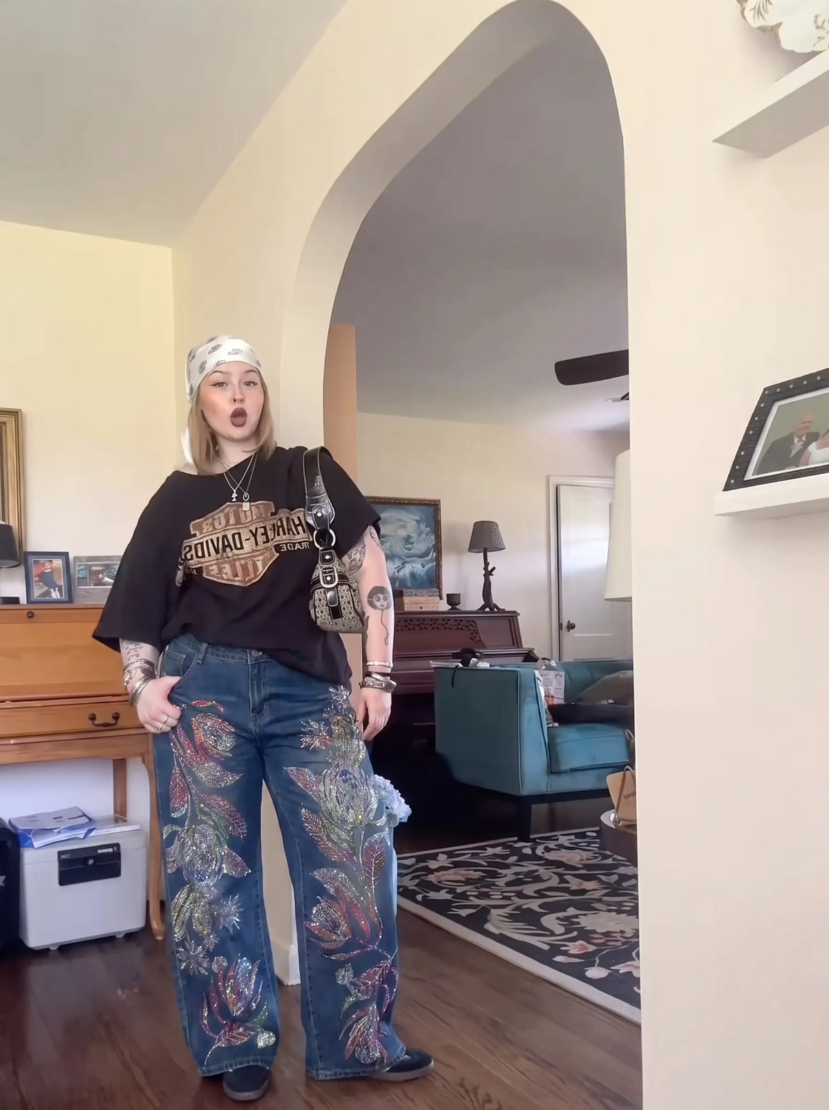 A person stands indoors wearing a black graphic T-shirt with a large vintage-style logo, wide-leg blue jeans with colorful floral embroidery, a white headband, and a small crossbody bag. The photo is taken in a home interior with an arched doorway, furniture, and decor visible in the background. TCH Floral Print Wide-Leg Denim Jeans