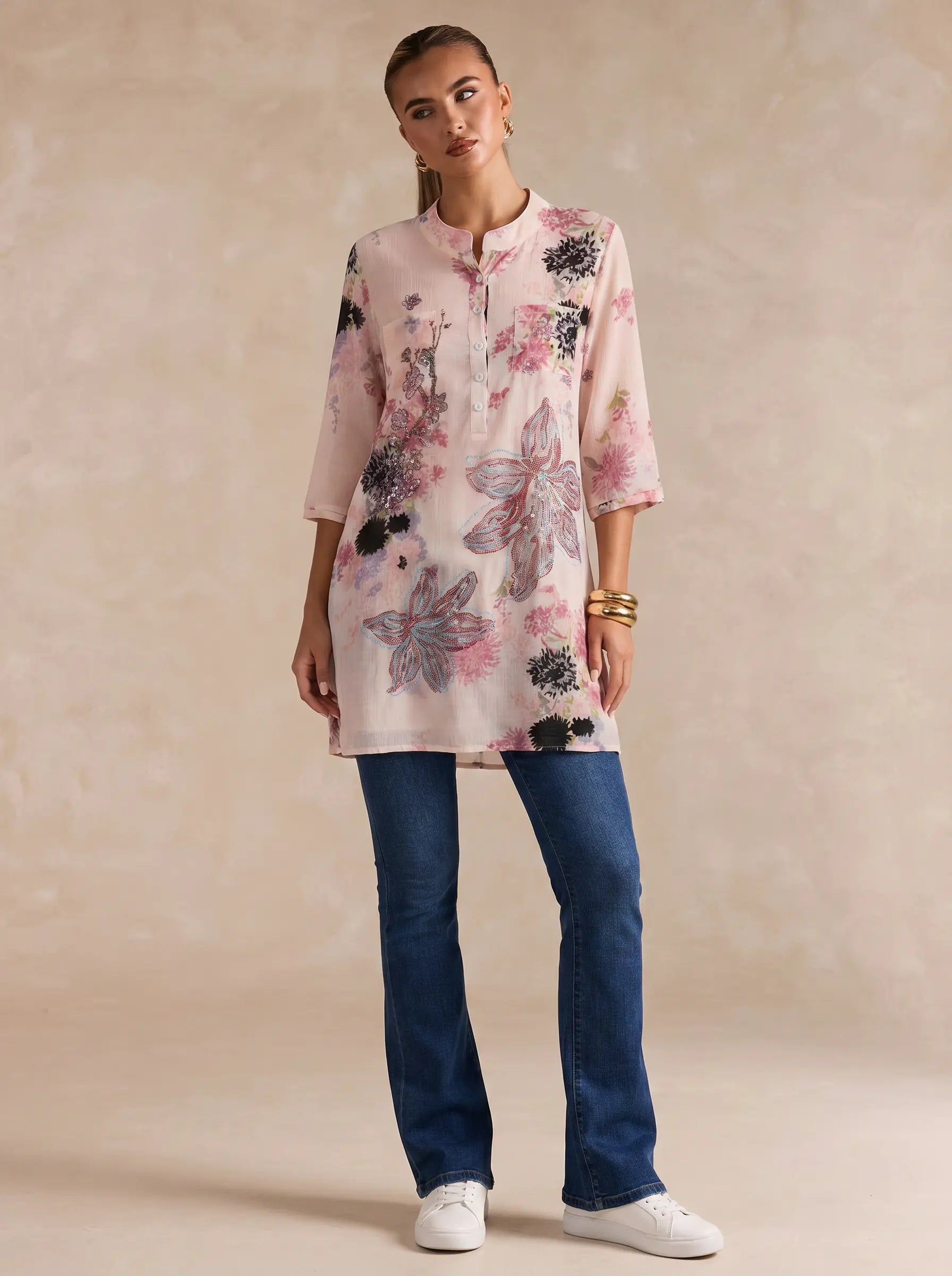 A woman modeling a light pink floral tunic top with blue jeans and white sneakers, standing against a textured beige studio backdrop. TCH Floral Rhinestone Butterfly Print Short Sleeve Blouse