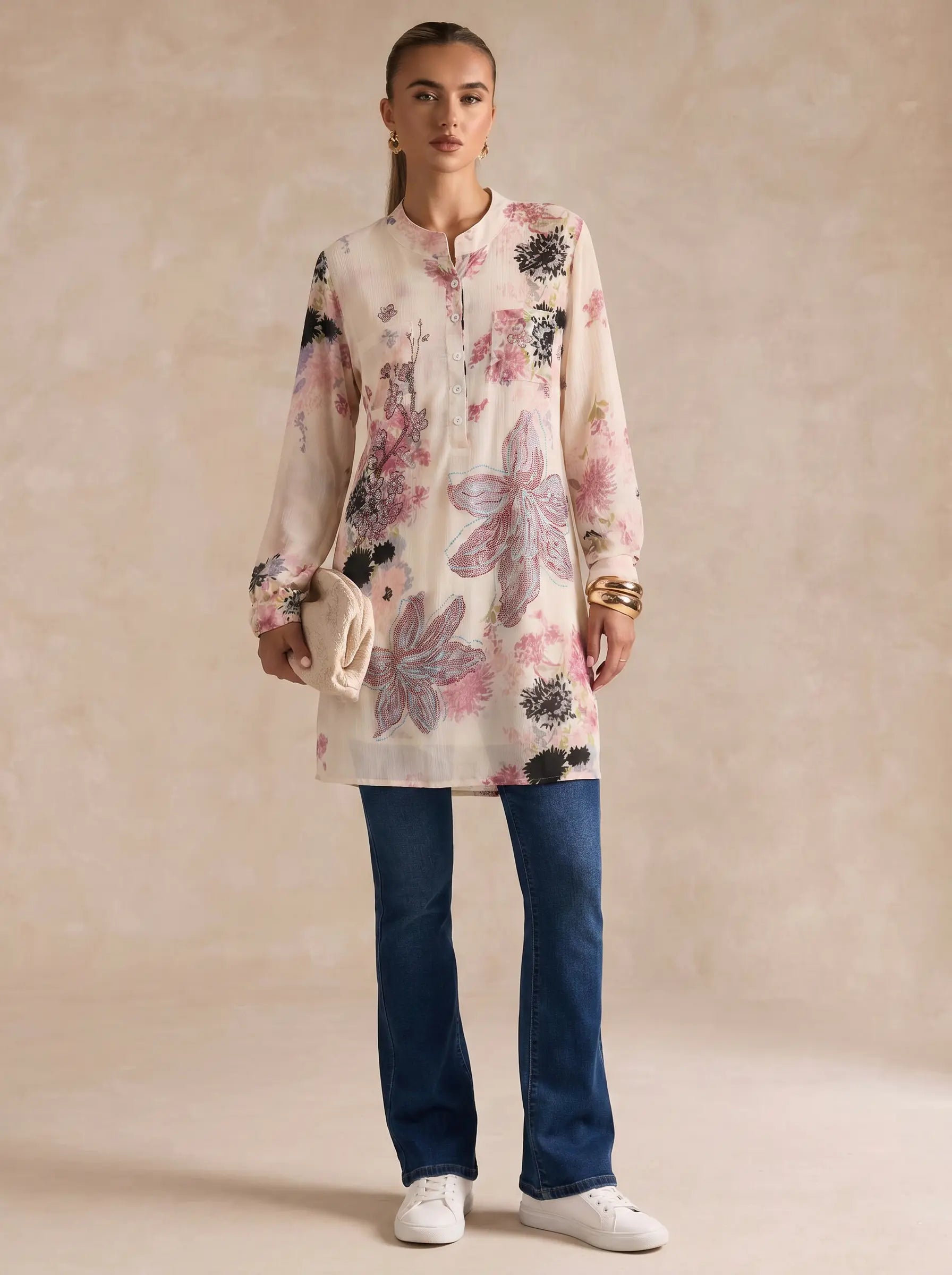 A woman stands against a warm beige studio backdrop wearing a long-sleeve floral tunic top with blue jeans and white sneakers, holding a small cream clutch. TCH Floral Rhinestone Butterfly Print Short Sleeve Blouse